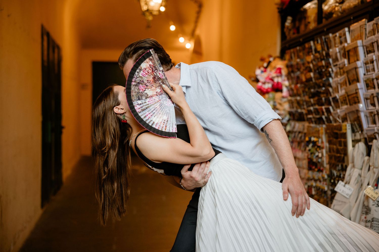 A romantic couple shares an intimate moment in a warmly lit indoor hallway with decorative lights in the background.