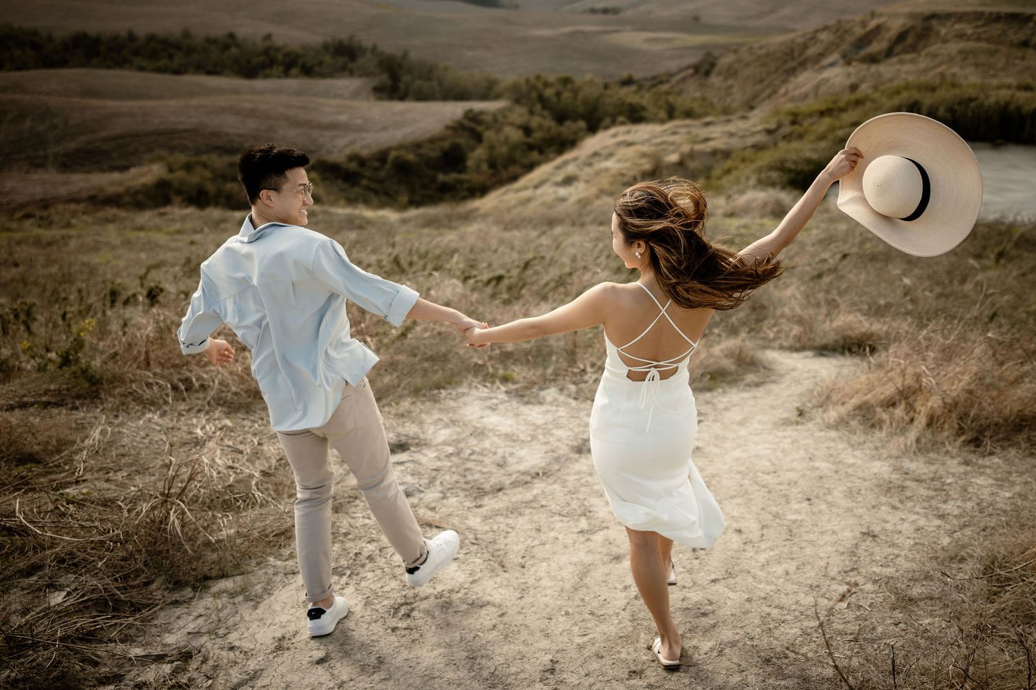 A couple in white outfits dancing playfully on a hilltop with a straw hat flying in the wind.