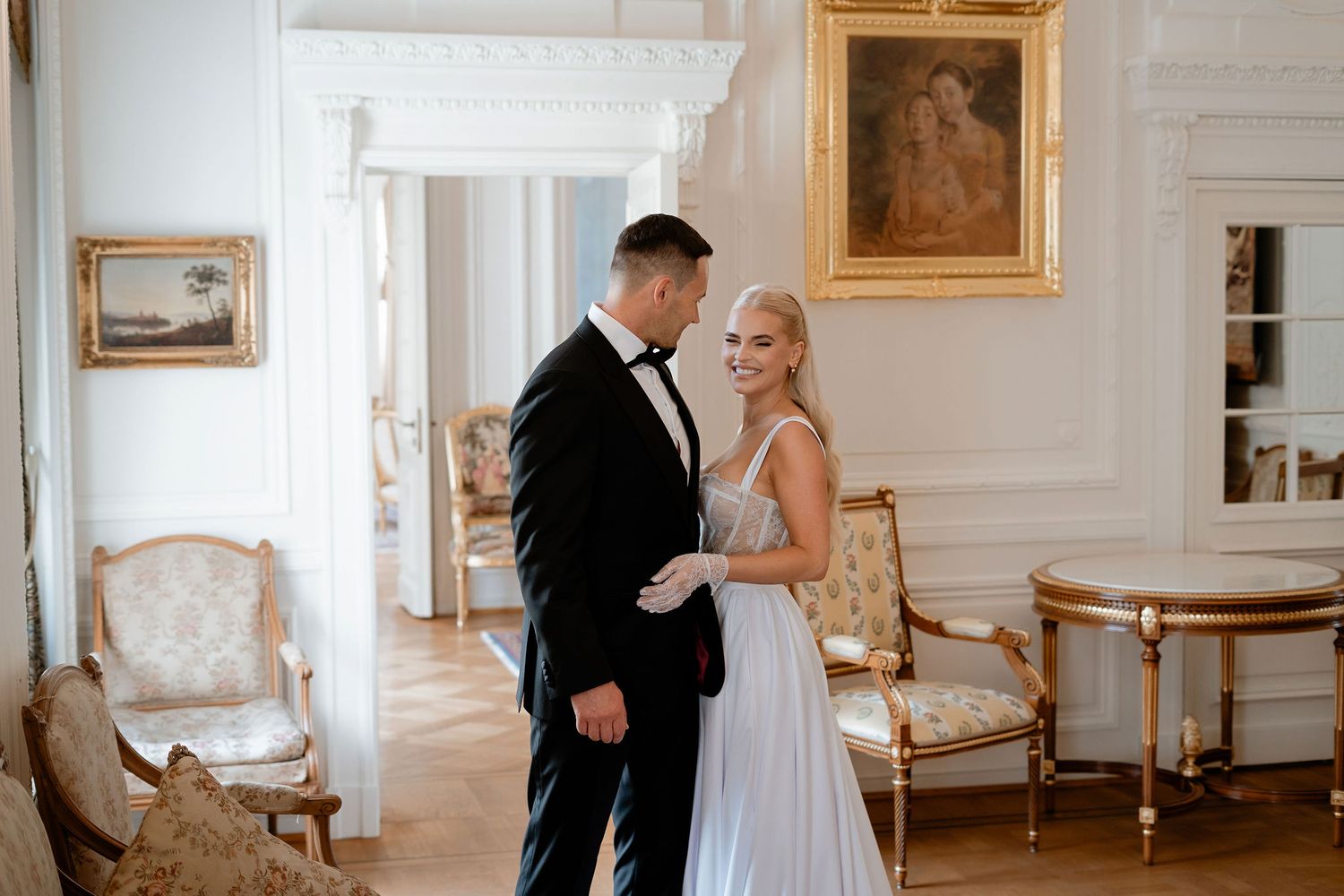 Elegant couple poses in vintage-style room with antique furniture and artwork.