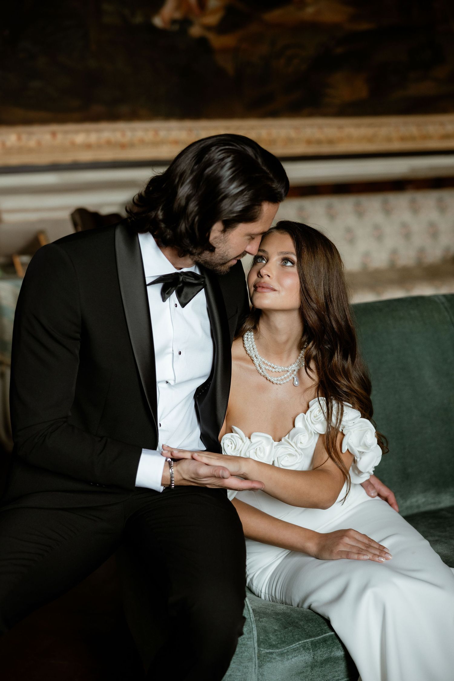 Elegant bride and groom share an intimate moment on a vintage green sofa in formal wedding attire.
