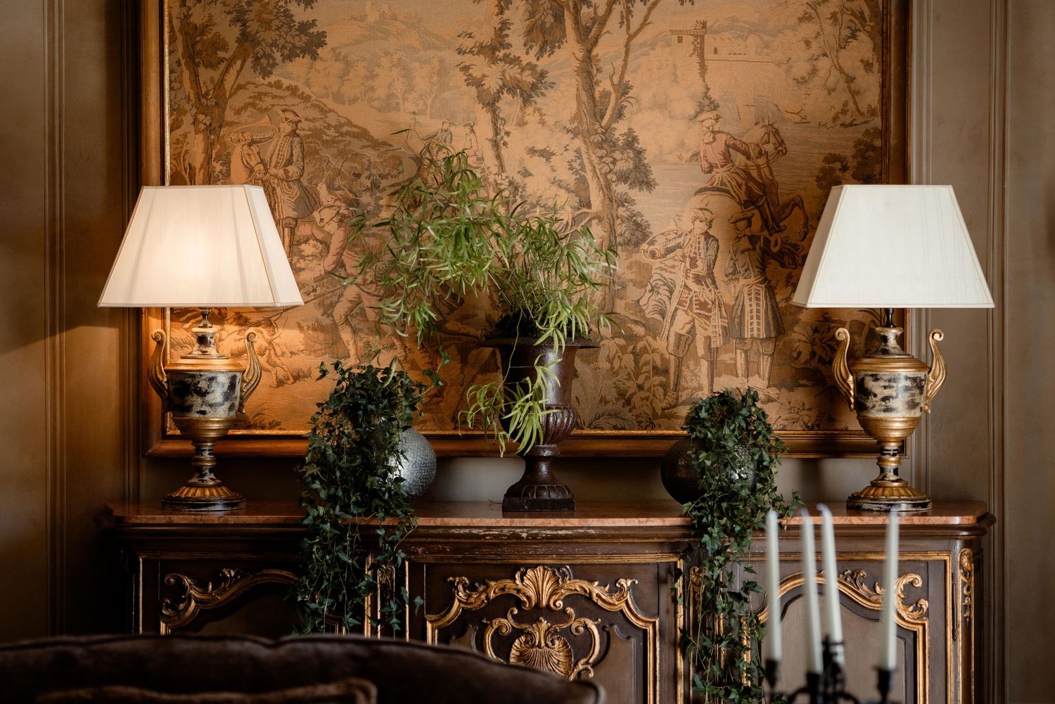 Elegant antique sideboard with matching table lamps and decorative greenery against a textured wallpaper background.
