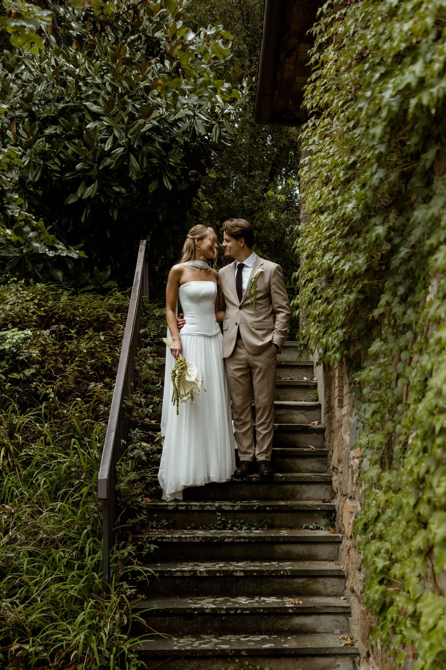 A couple in wedding attire walking up rustic wooden stairs surrounded by lush green foliage in a garden setting.