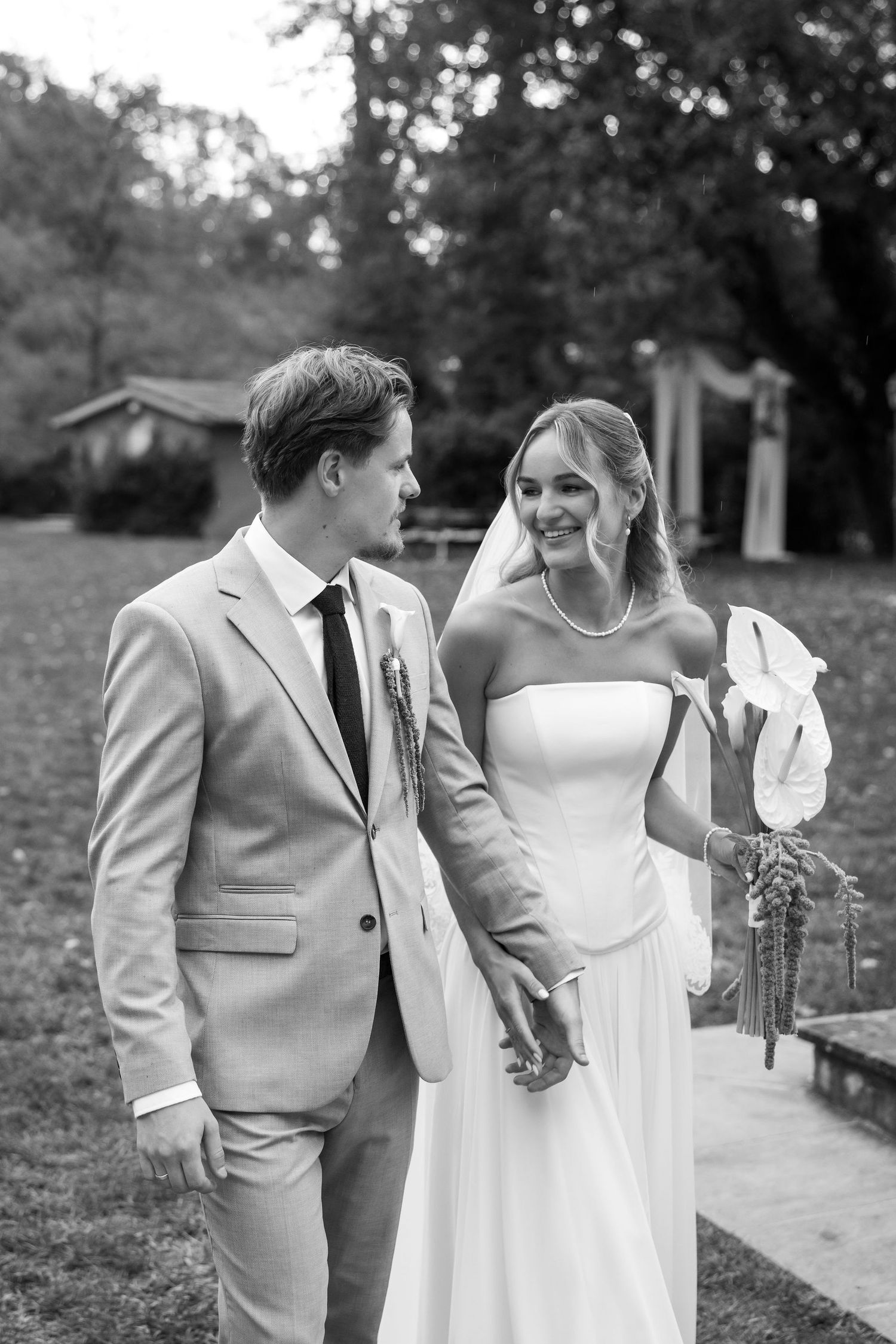 A couple in wedding attire share a candid moment together in a garden setting, captured in black and white.