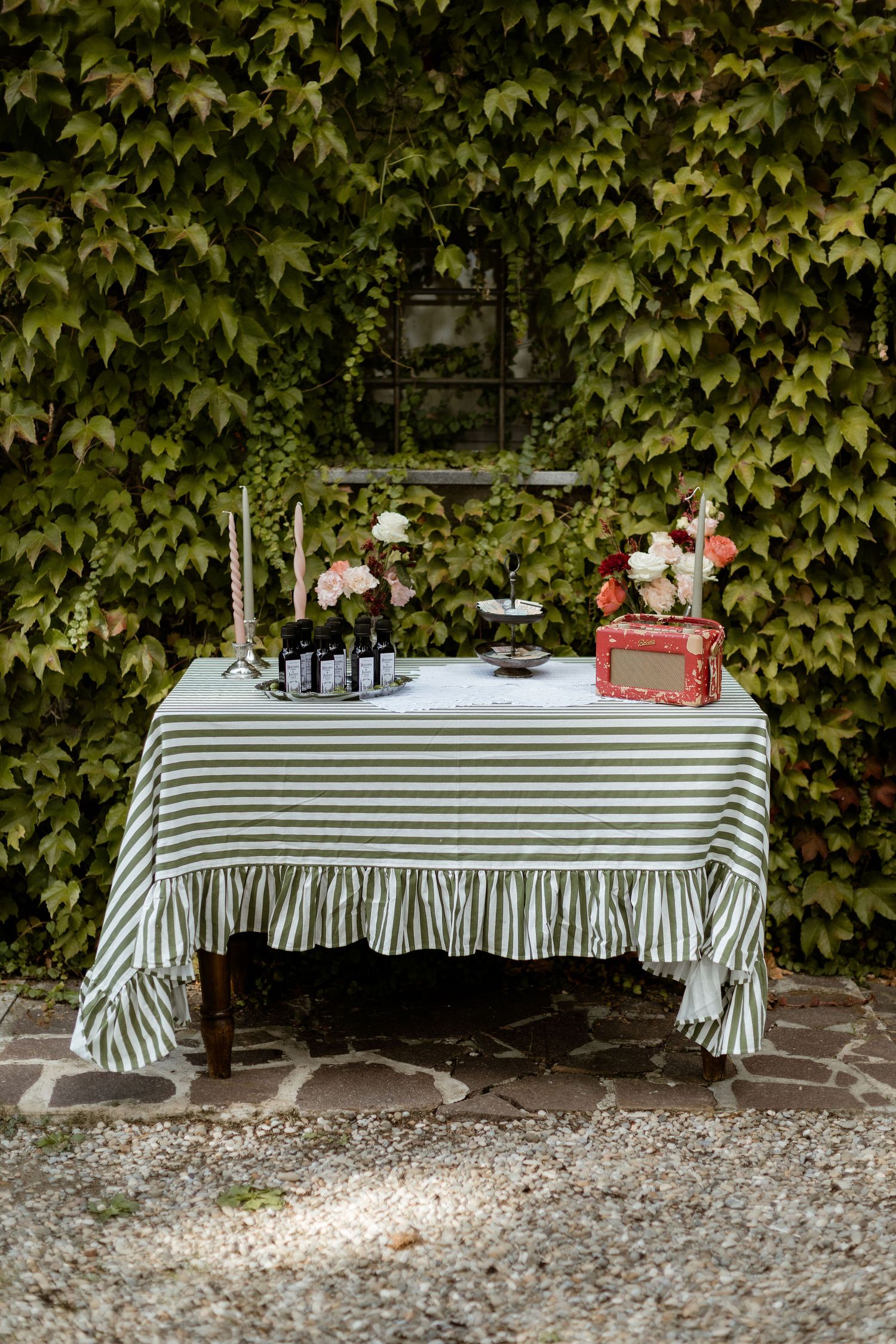 A series of tables with striped tablecloths and decorative items arranged against a vine-covered wall in a garden setting.