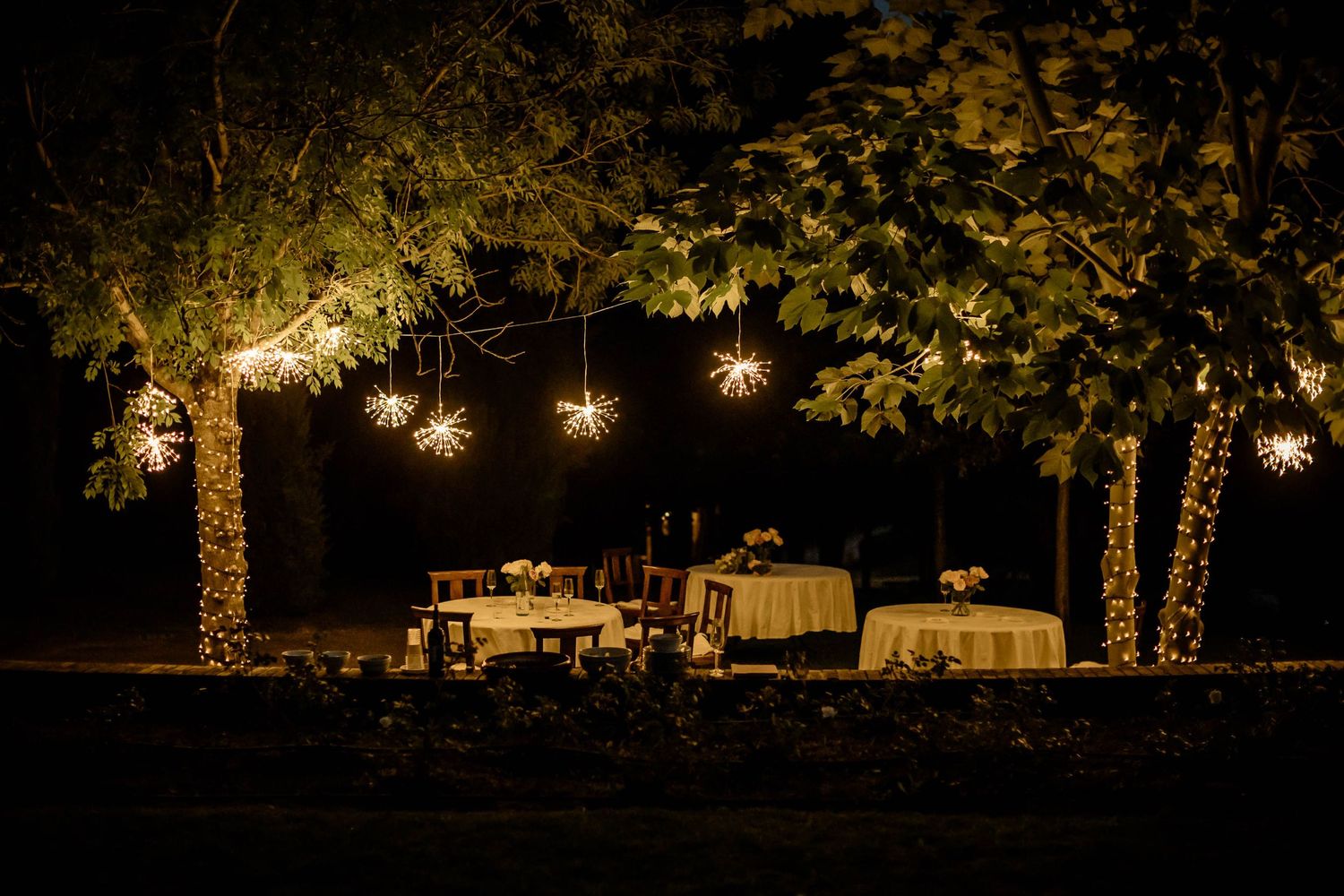 Outdoor evening wedding reception with hanging lights and decorated tables under trees.