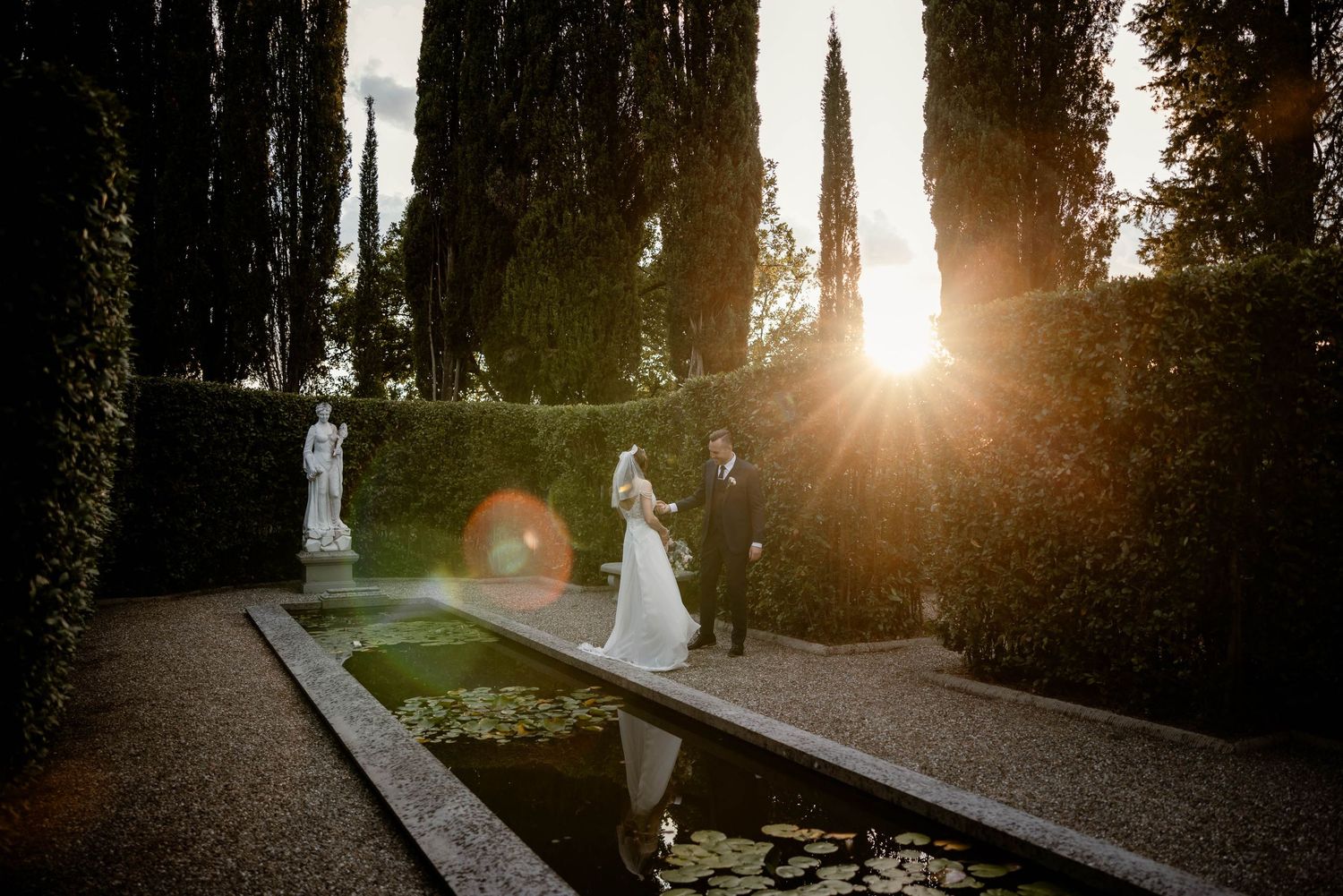 Sunset wedding photo in Italian garden with cypress trees creating dramatic silhouettes.