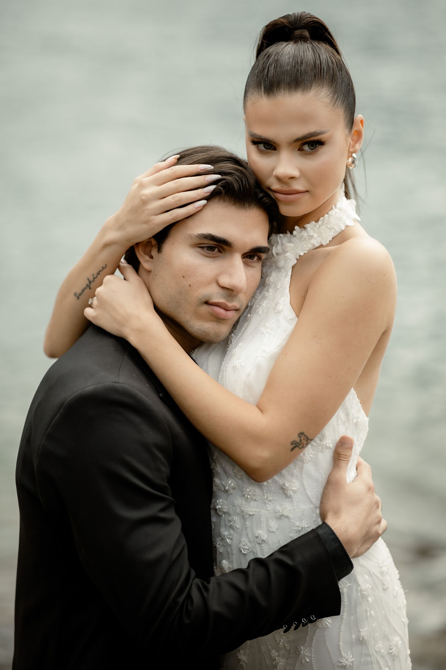 Elegant couple in formal attire embrace against misty waterfront backdrop.