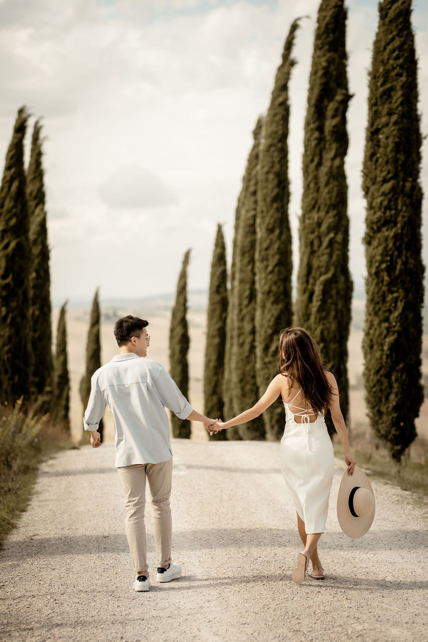 Couple walking hand in hand down iconic Italian cypress tree road.