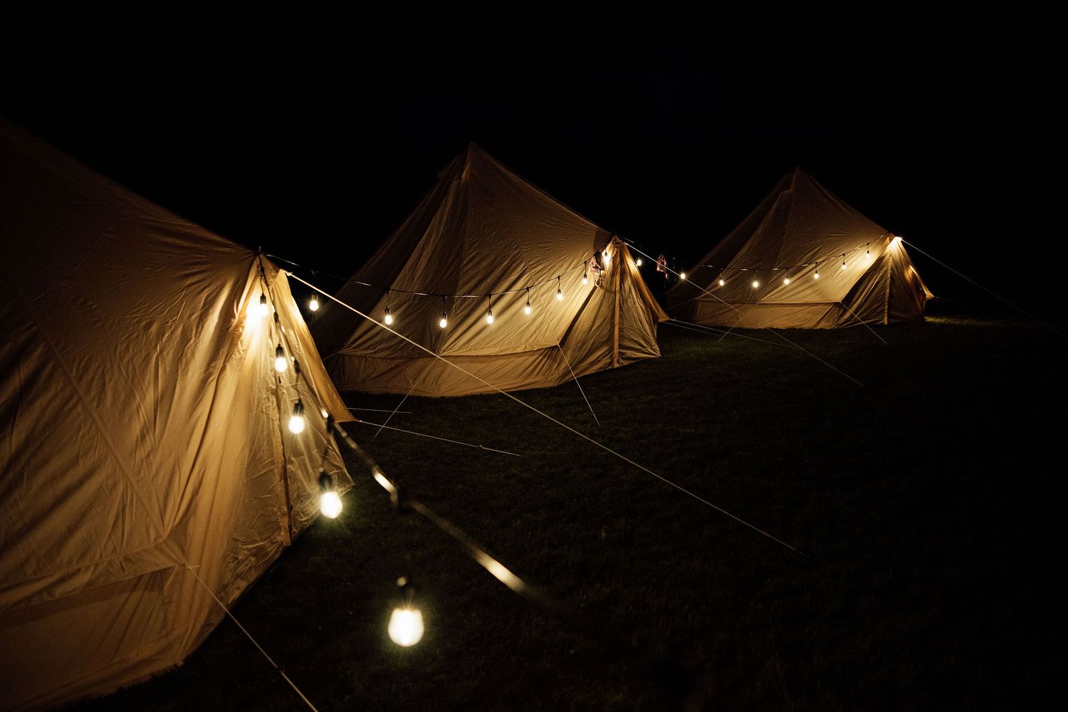 Canvas bell tents illuminated by string lights create a cozy nighttime glamping atmosphere in the darkness.