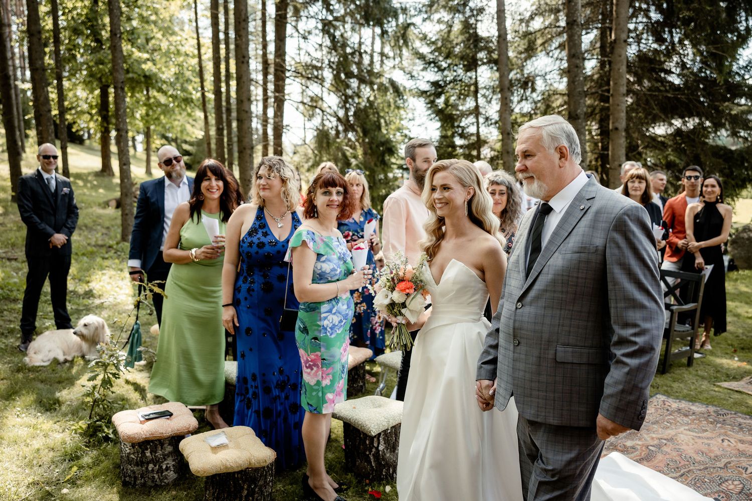 Wedding ceremony guests gather in wooded outdoor setting with rustic seating.