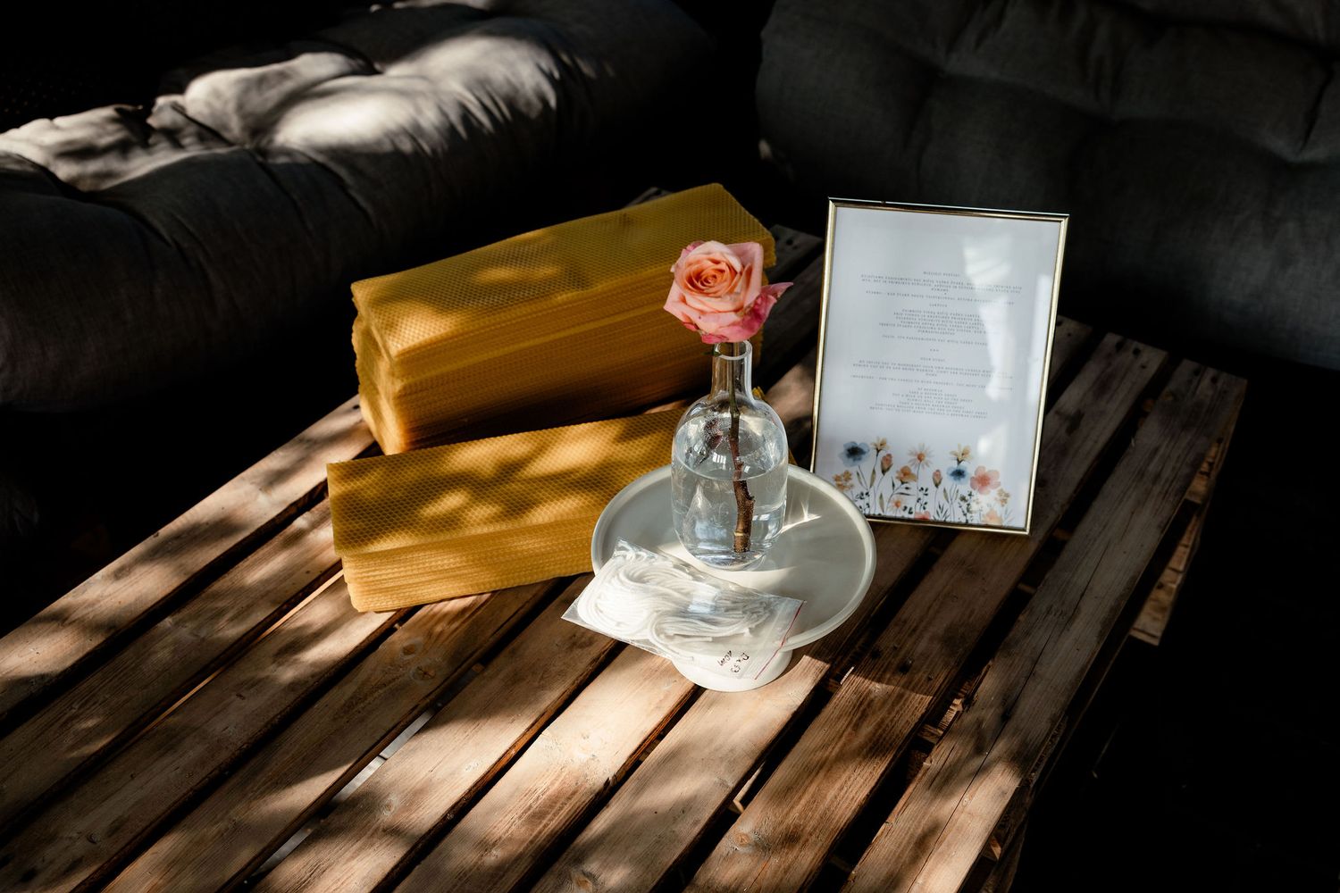 A single rose in a vase sits beside a frame on a wooden table with dramatic lighting.
