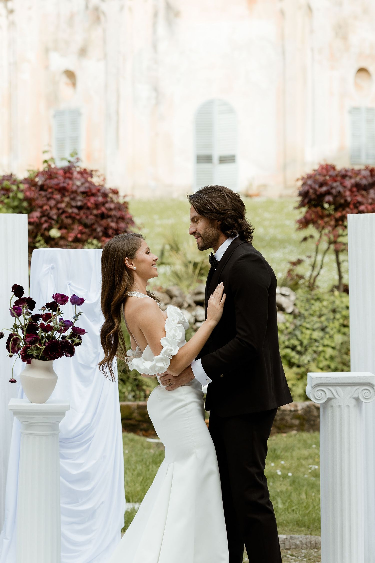 A romantic wedding ceremony outdoors with white pillars and burgundy florals decorating an elegant garden setting.