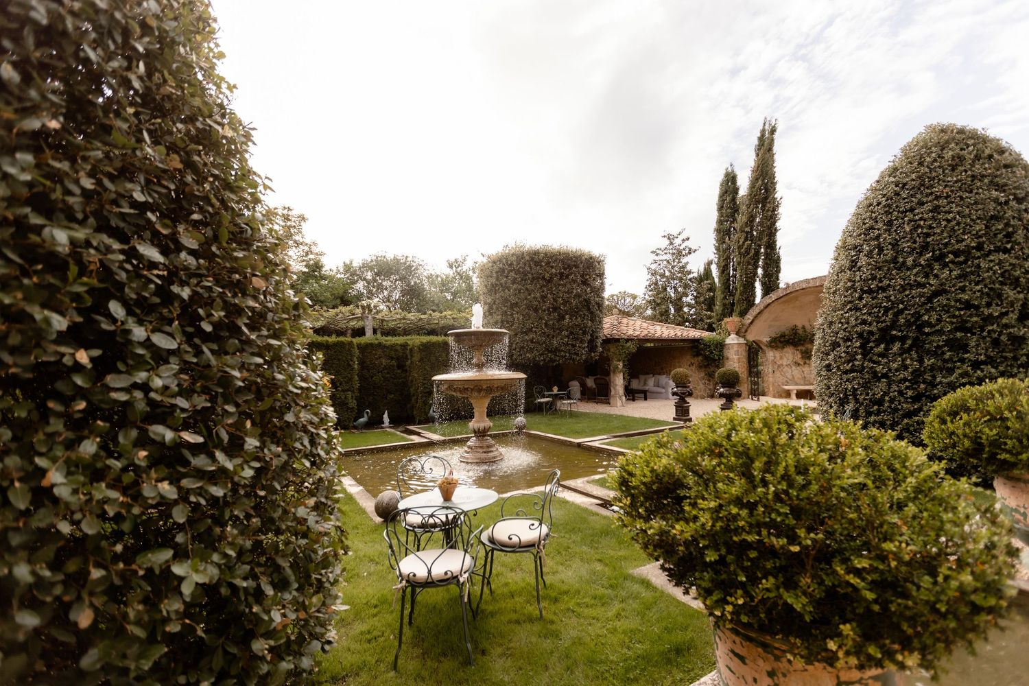 Garden patio with white tables and chairs surrounded by neatly trimmed hedges and cypress trees in an outdoor setting.