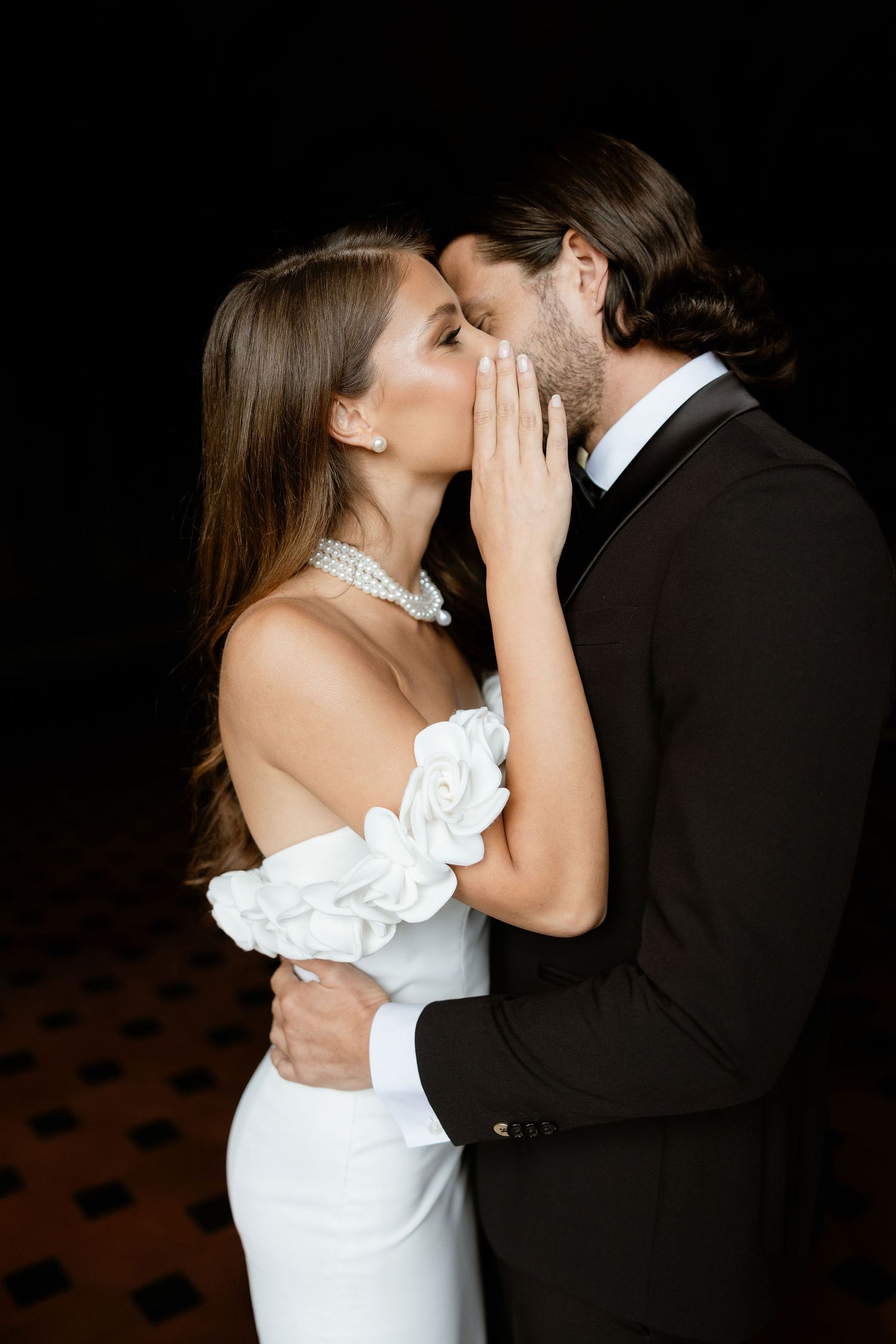 A couple shares an intimate moment in formal attire against a dark background with dramatic lighting.