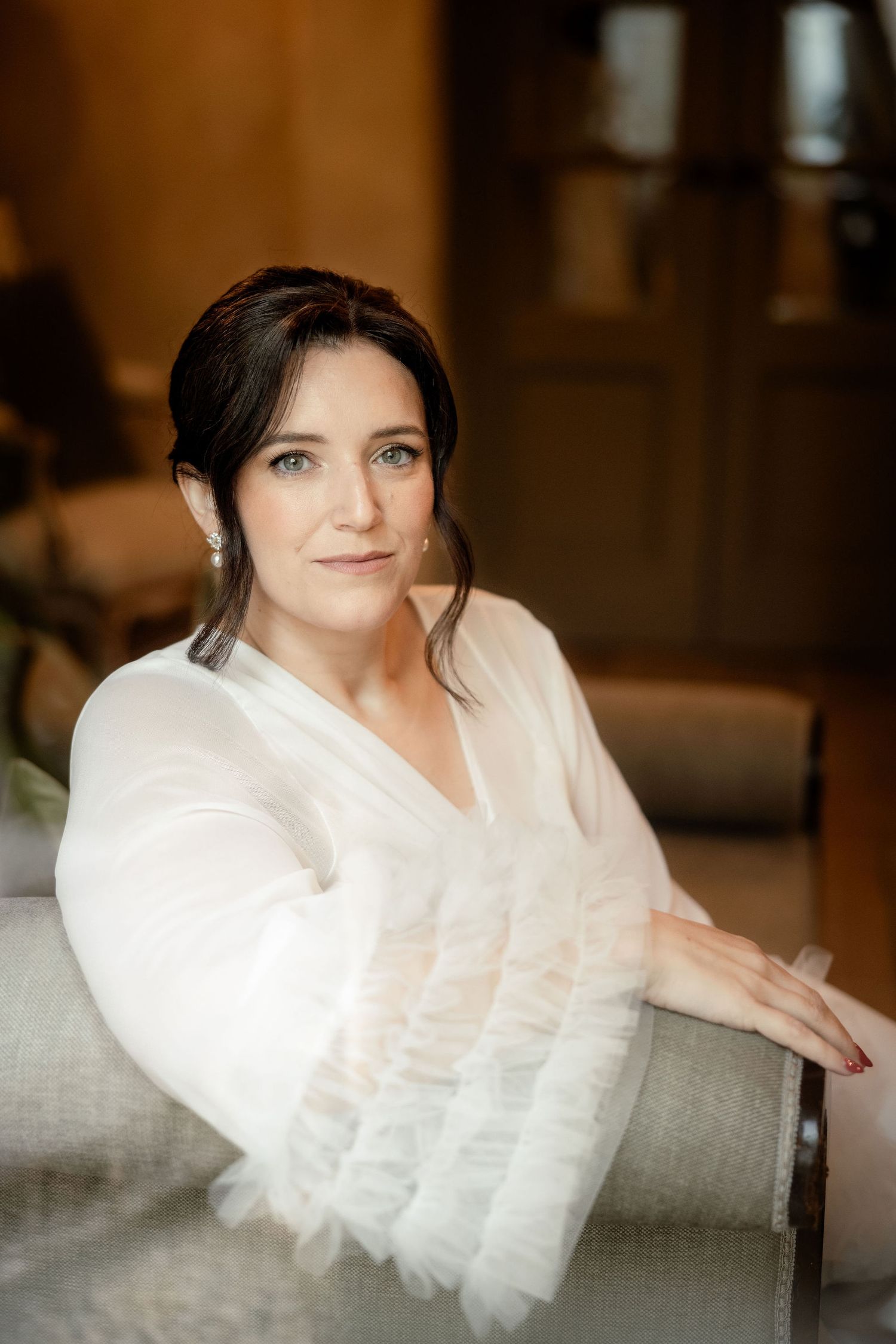 Portrait of person in white dress sitting on light gray couch in warm indoor setting with soft lighting.