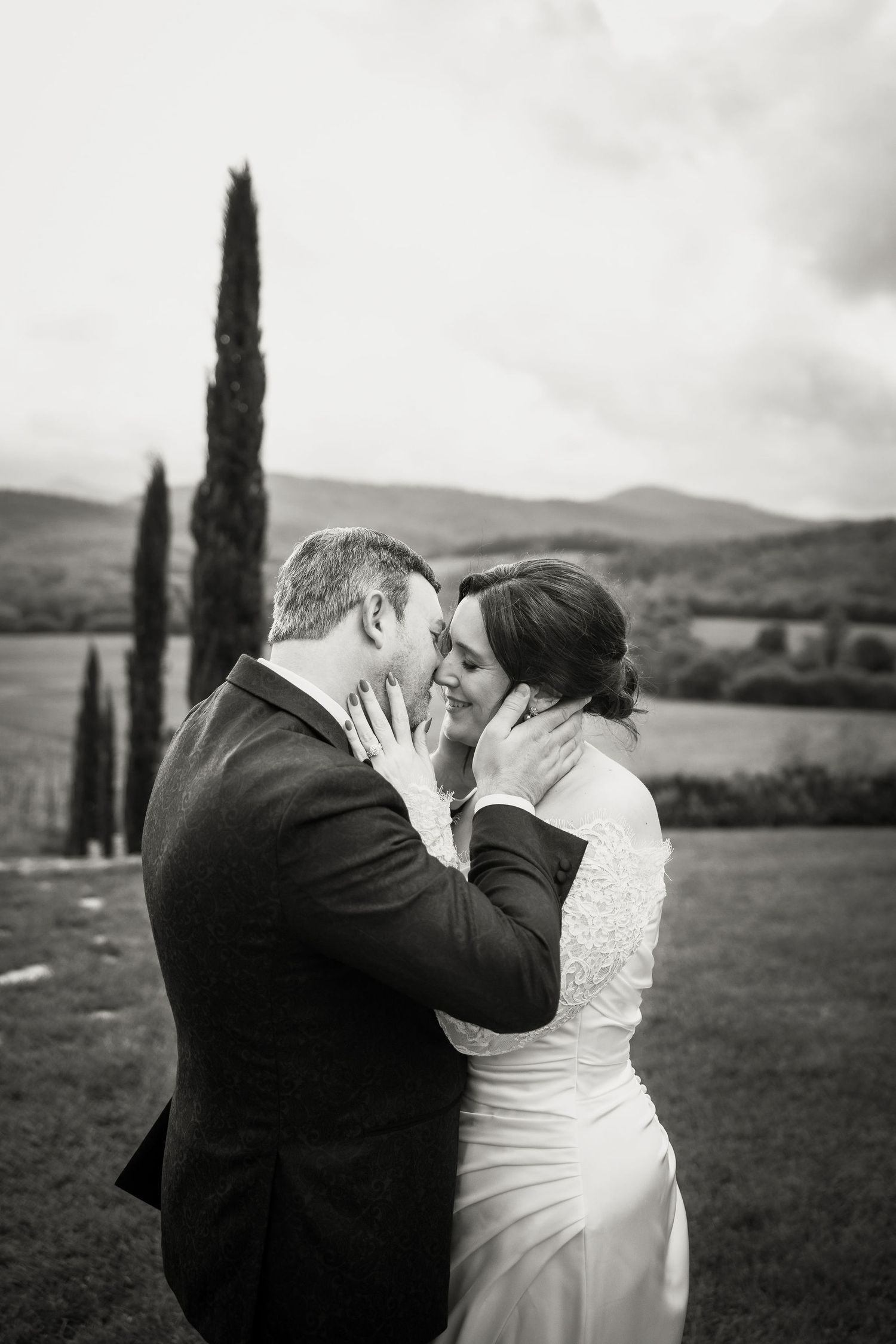 A romantic black and white wedding portrait shows a tender moment between a couple in front of cypress trees in Tuscany.