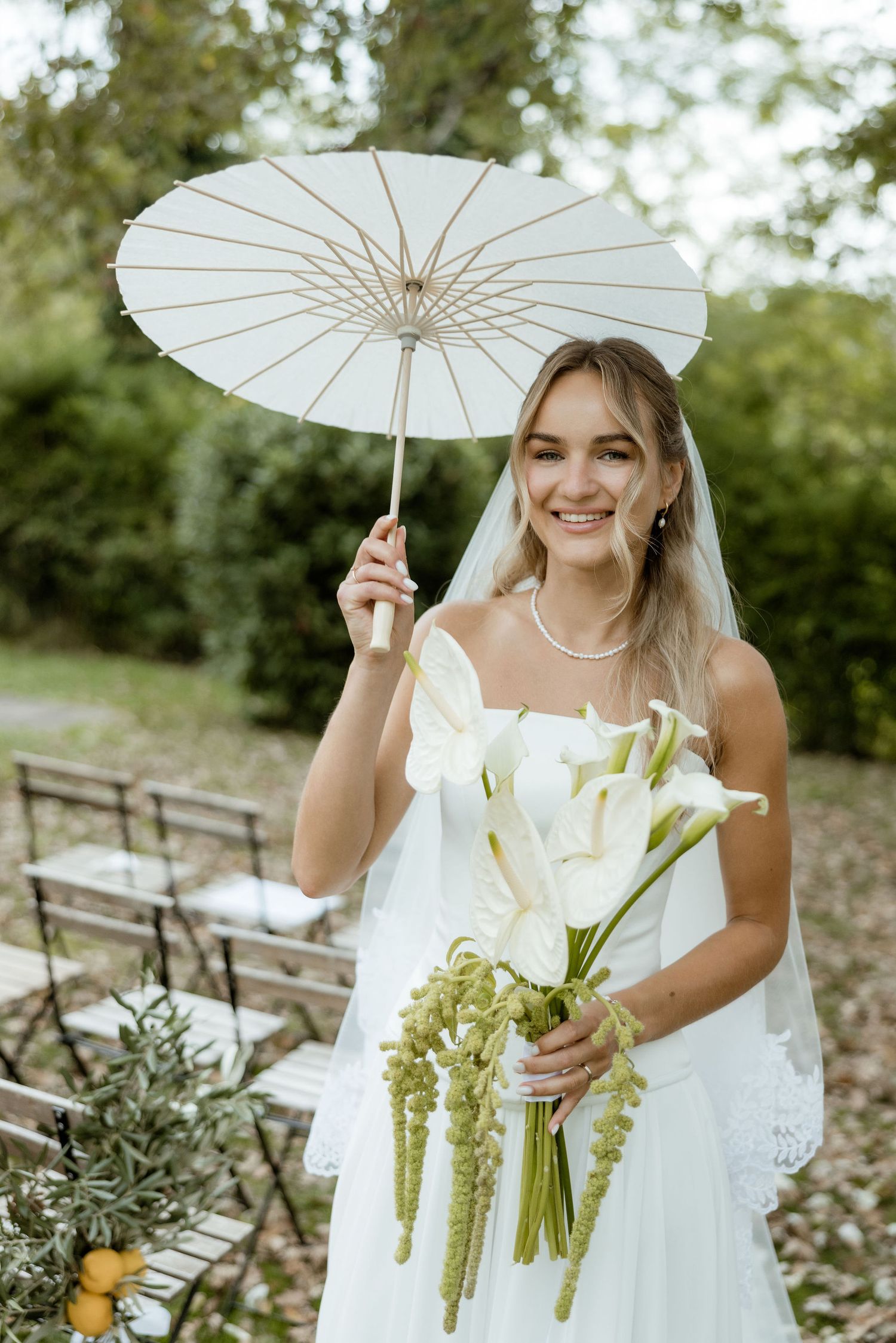 A bride in a white dress holds a delicate paper parasol and elegant white lily bouquet in an outdoor garden setting.