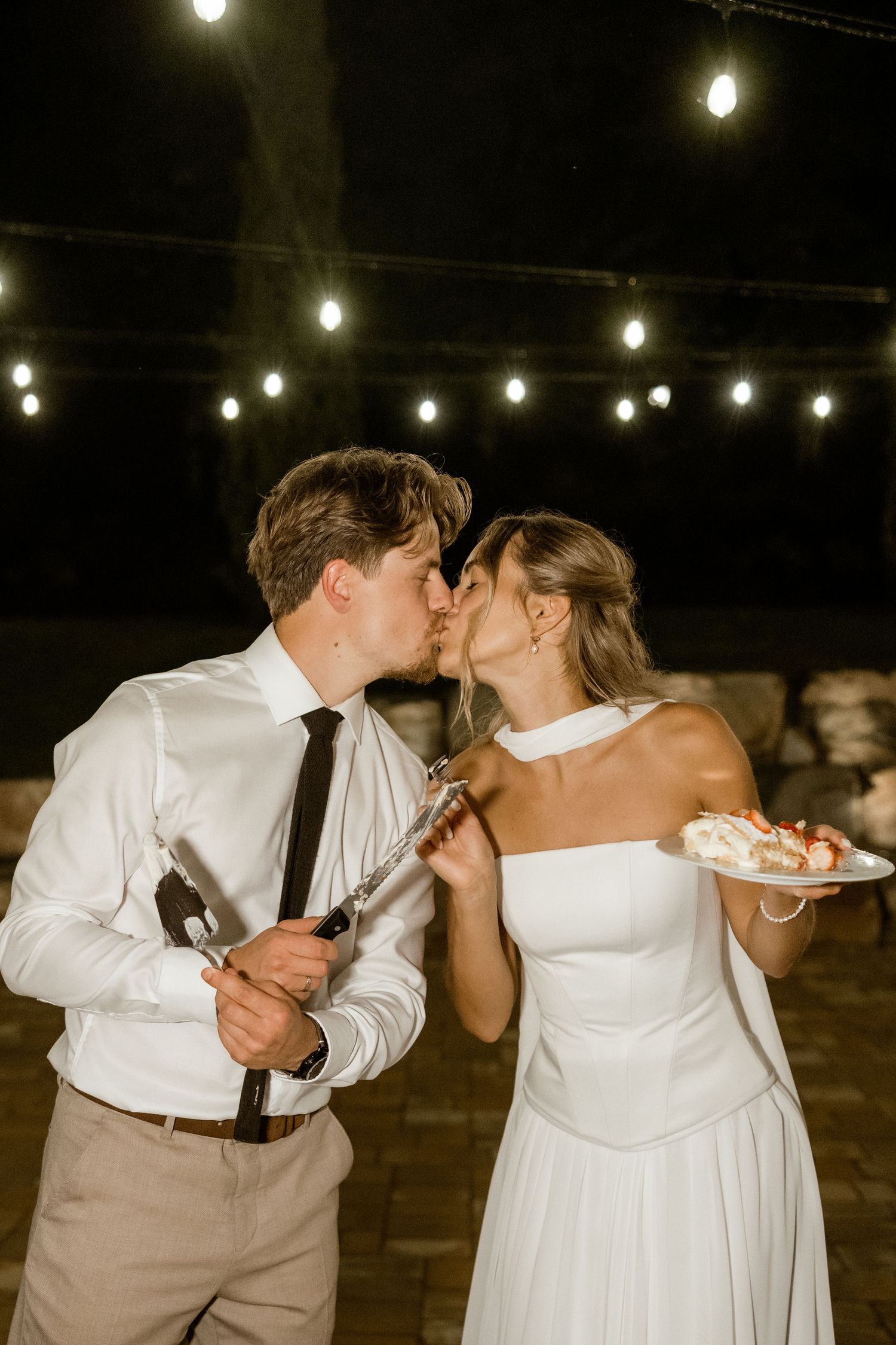 A romantic moment under string lights as newlyweds share dessert at their outdoor wedding reception.