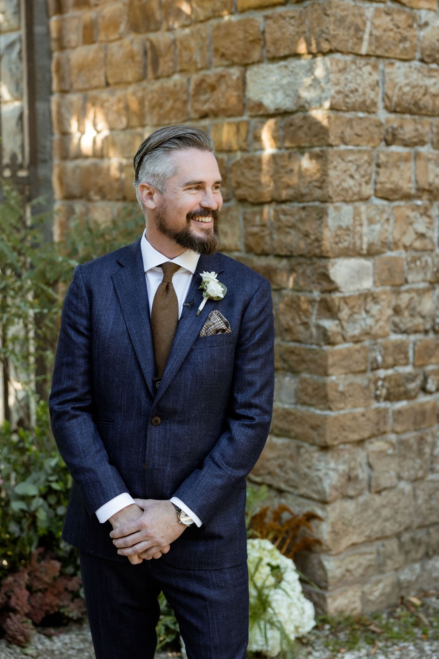 A groom in a navy pinstripe suit and brown tie stands against a stone wall background.