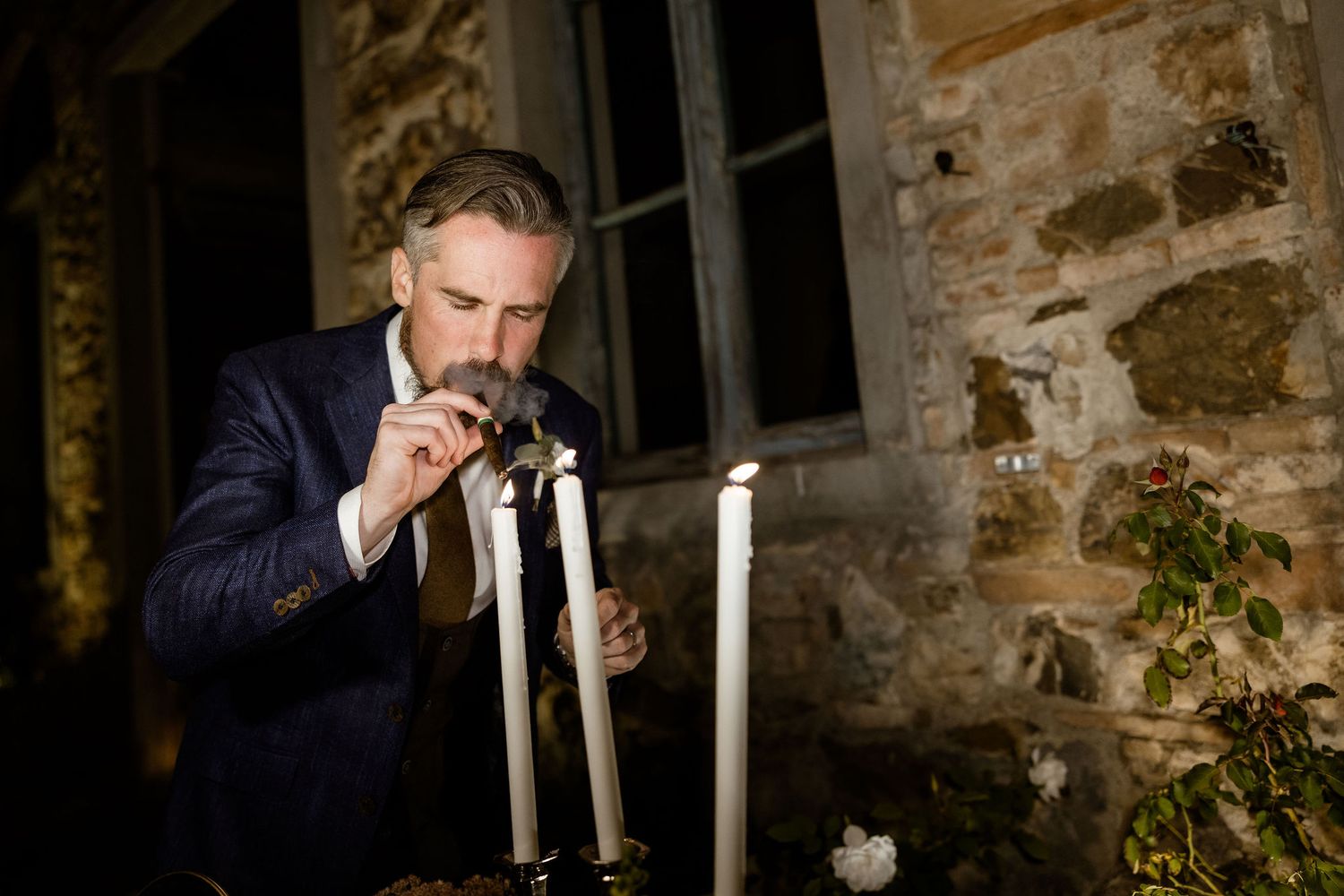 A person in a dark suit holds a glass in front of a rustic stone wall with candlelight ambiance.