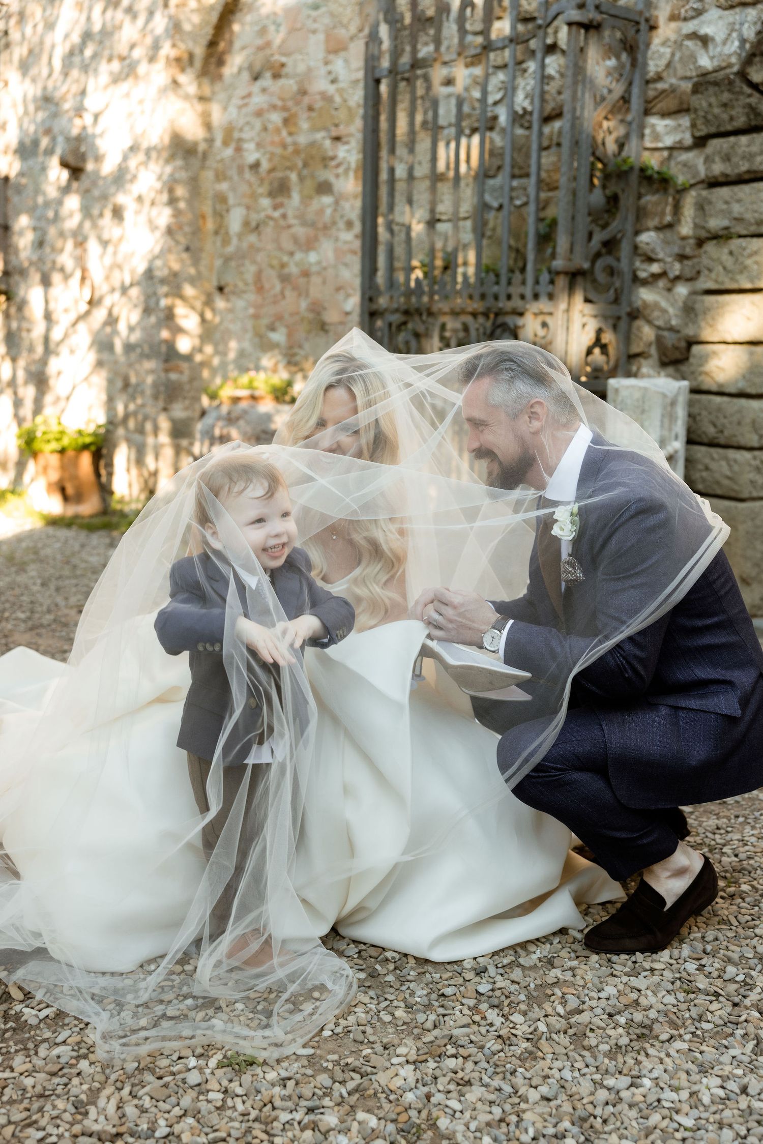 A romantic wedding couple shares tender moments under a flowing veil against a rustic stone wall backdrop.