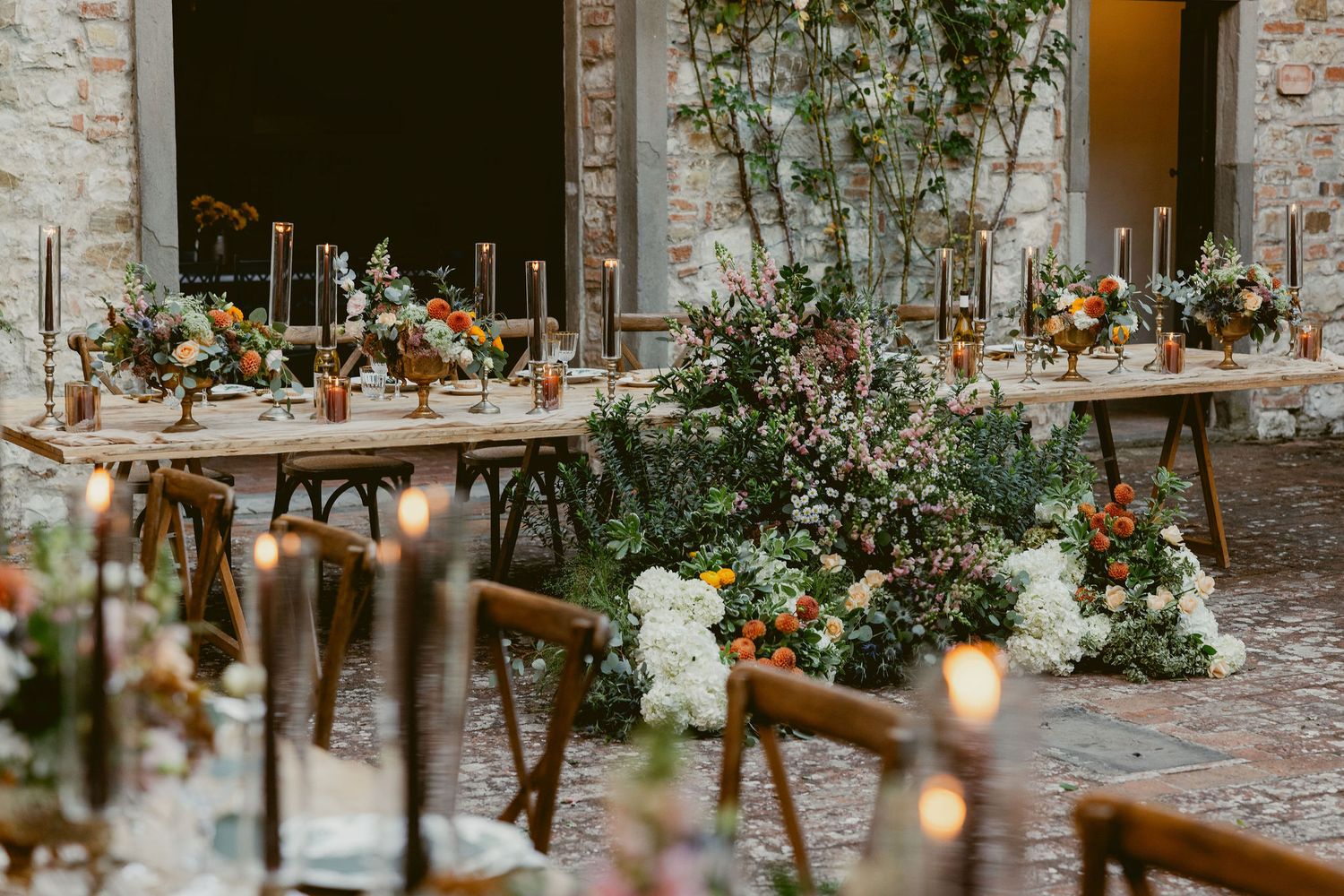 Elegant rustic wedding reception tables decorated with greenery, candles and floral arrangements in a stone-walled venue.