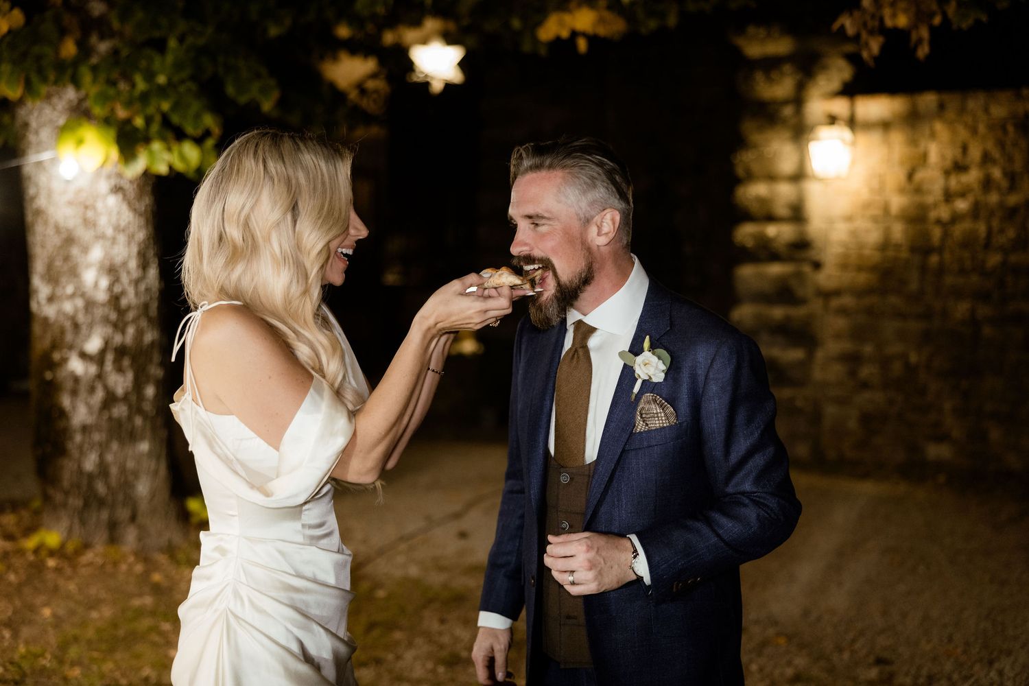 A couple shares intimate moments during their evening wedding photoshoot against a rustic brick wall backdrop.