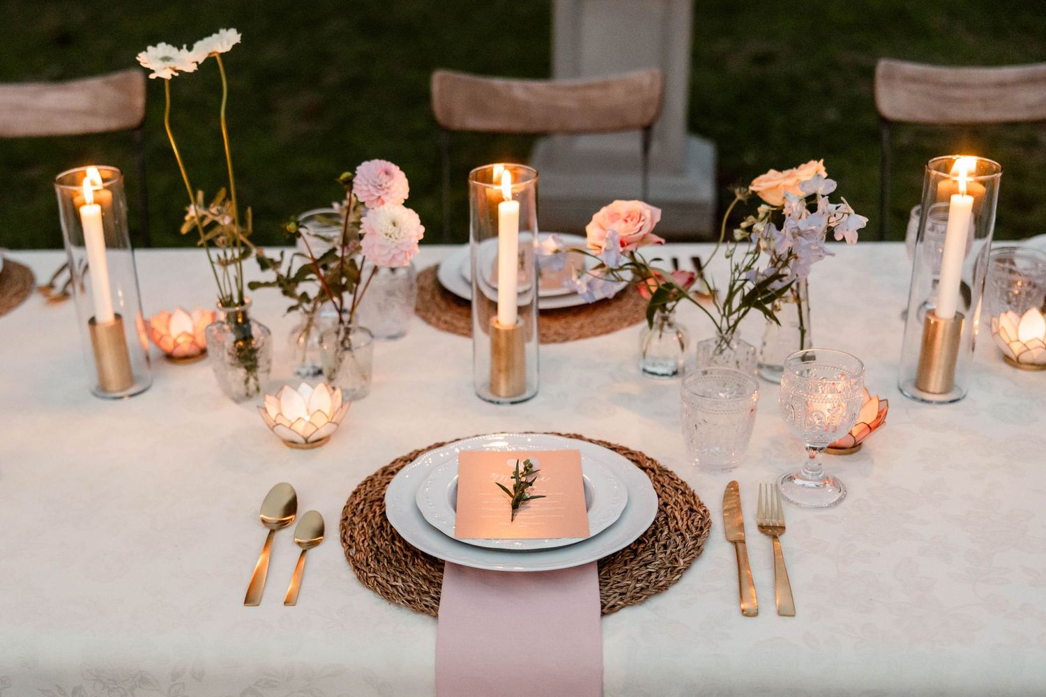 Romantic table setting with candles, pink flowers, and woven placemats for an outdoor wedding.