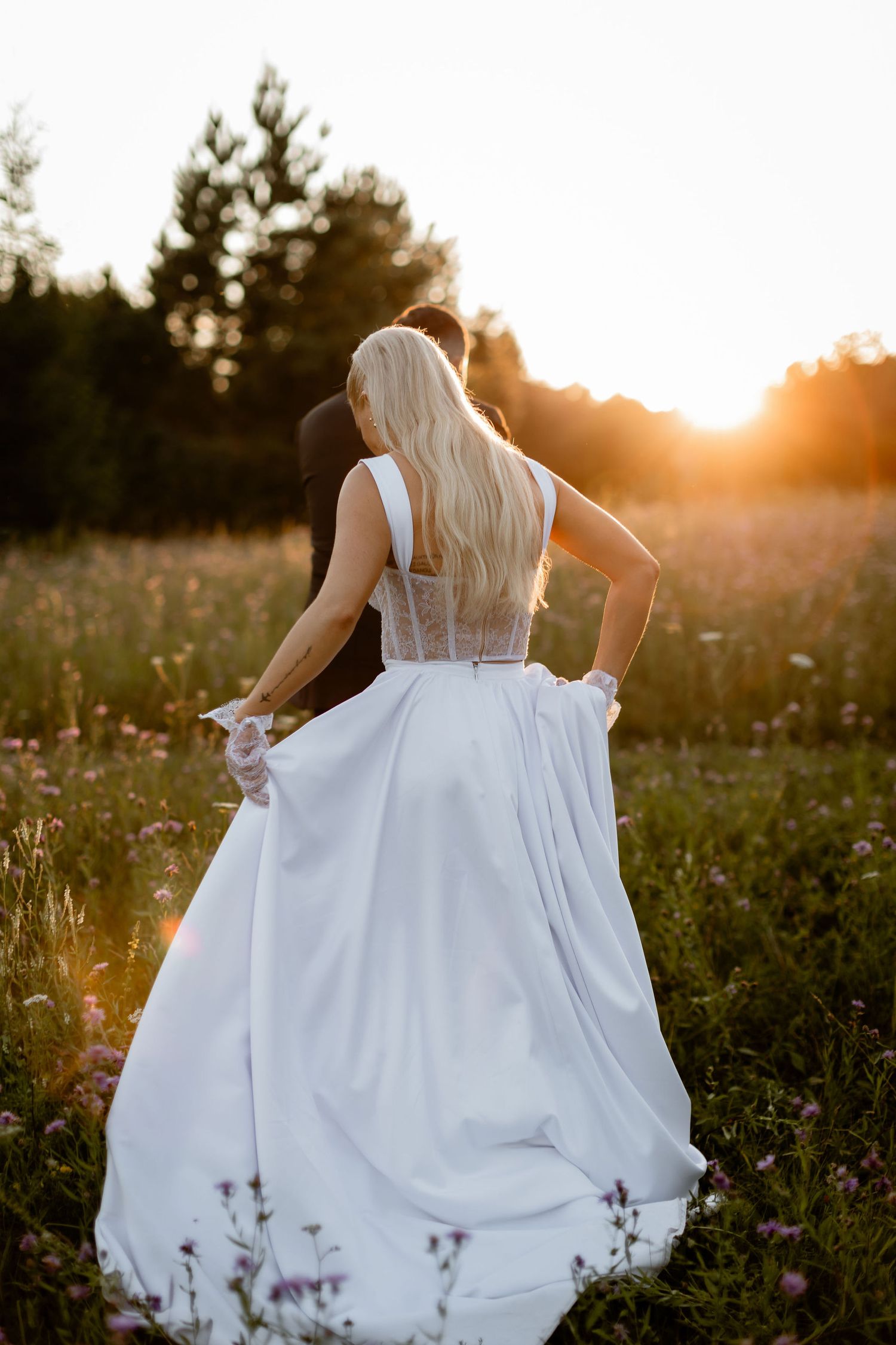 White wedding dress flowing in evening sunlight as bride stands in rustic meadow.