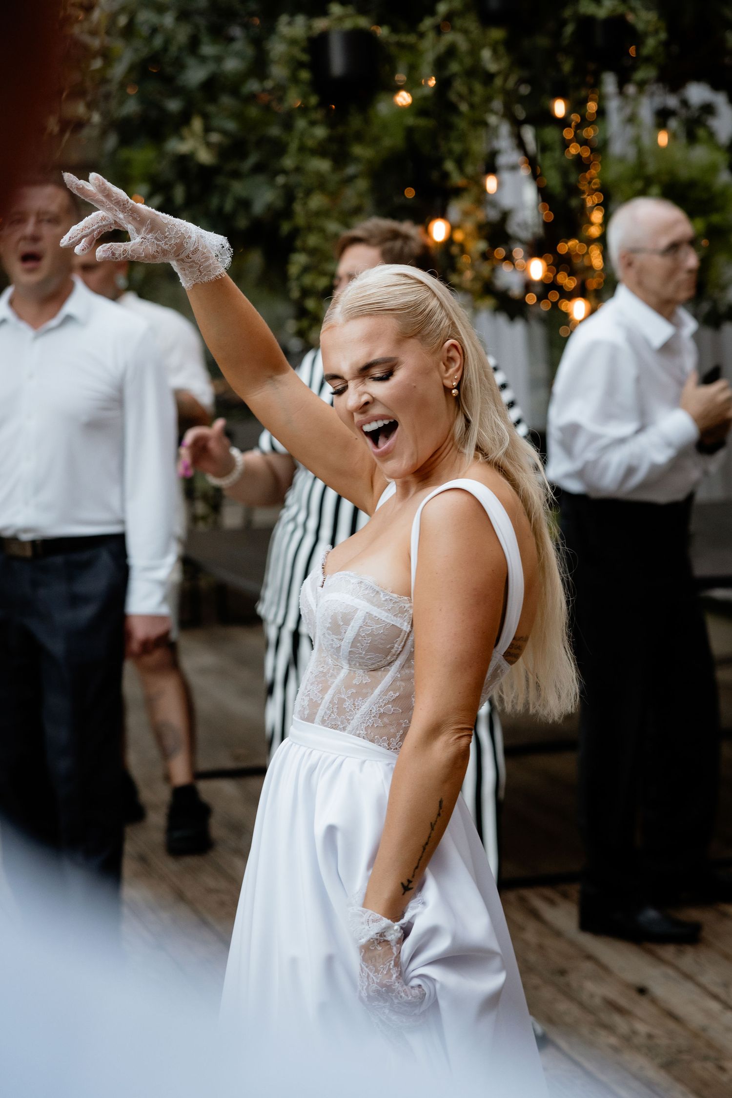 A bride in a white dress dances joyfully at an outdoor wedding reception with white gloves and flowing blonde hair.