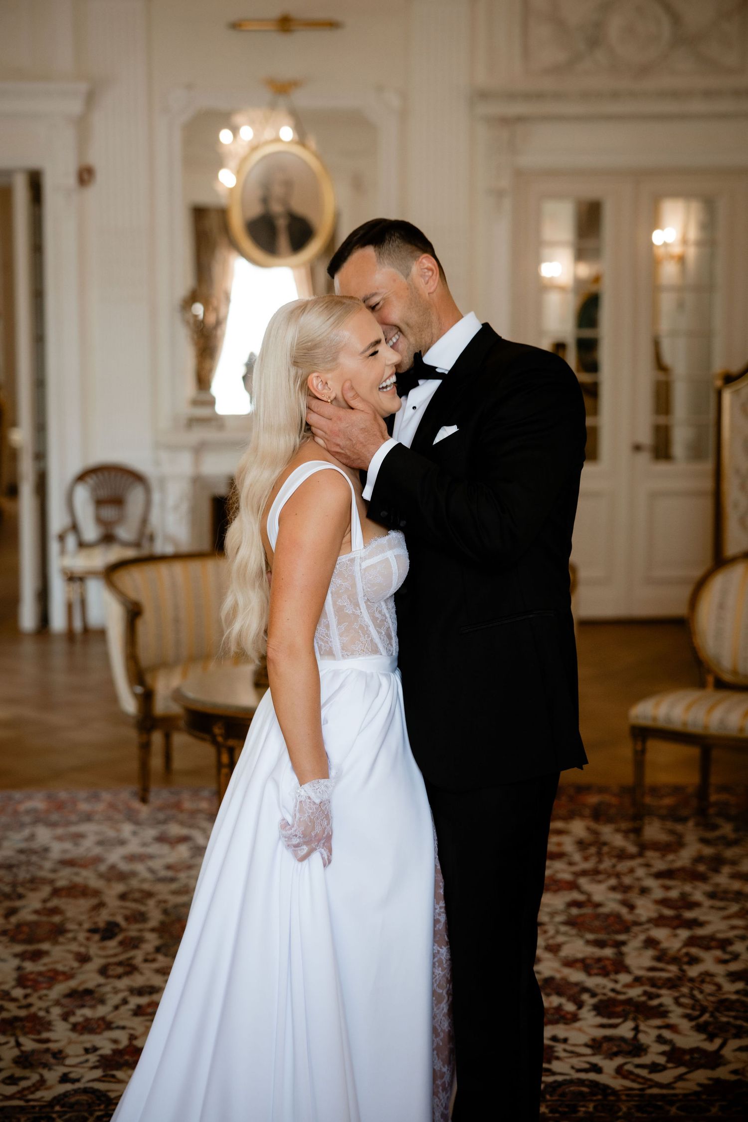 A romantic wedding couple embraces in an elegant ballroom with chandelier and vintage furnishings.