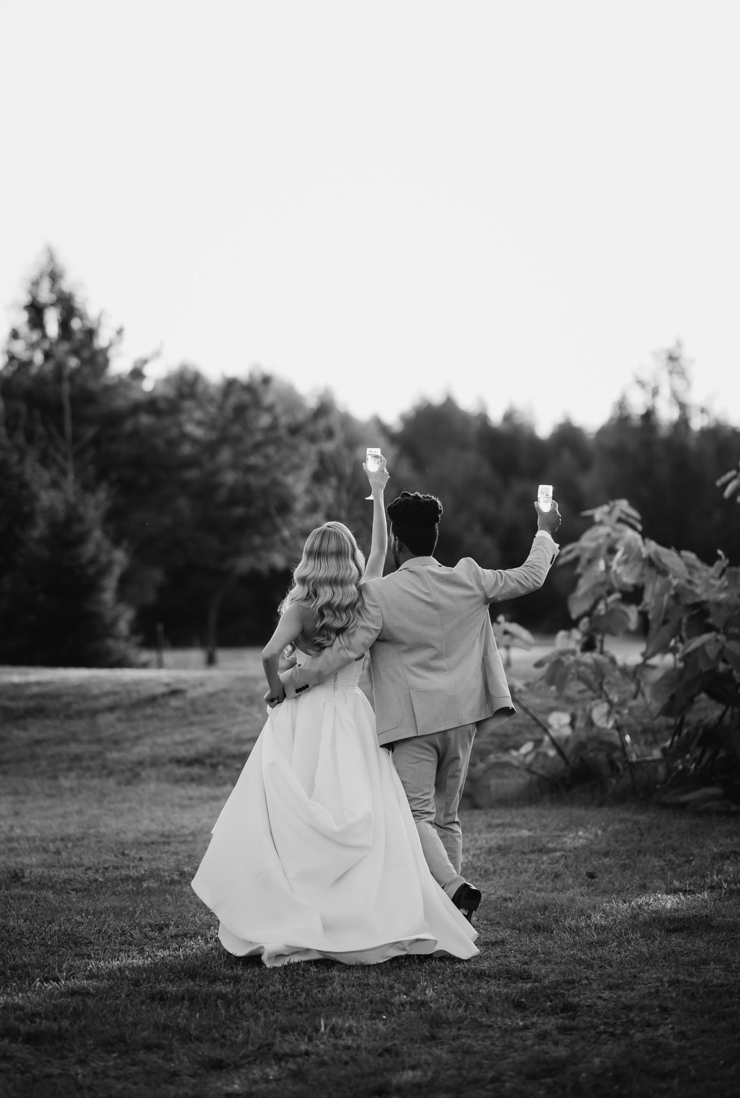 A couple in wedding attire joyfully dances together on a grassy lawn with trees in the background.