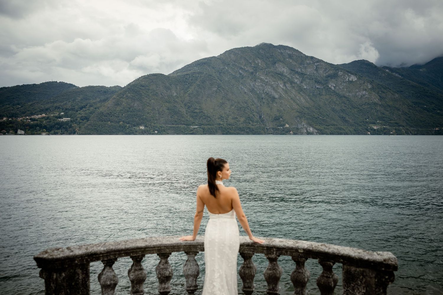 Bride in white dress looks out over mountain lake from stone balcony terrace.