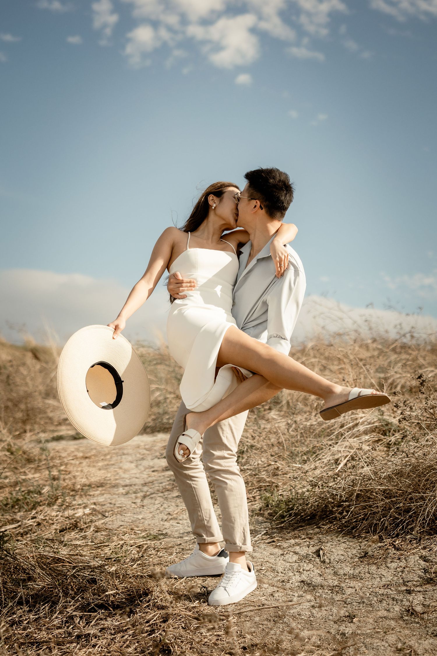 A romantic embrace on a hillside with a straw hat and white summer outfits against blue skies.
