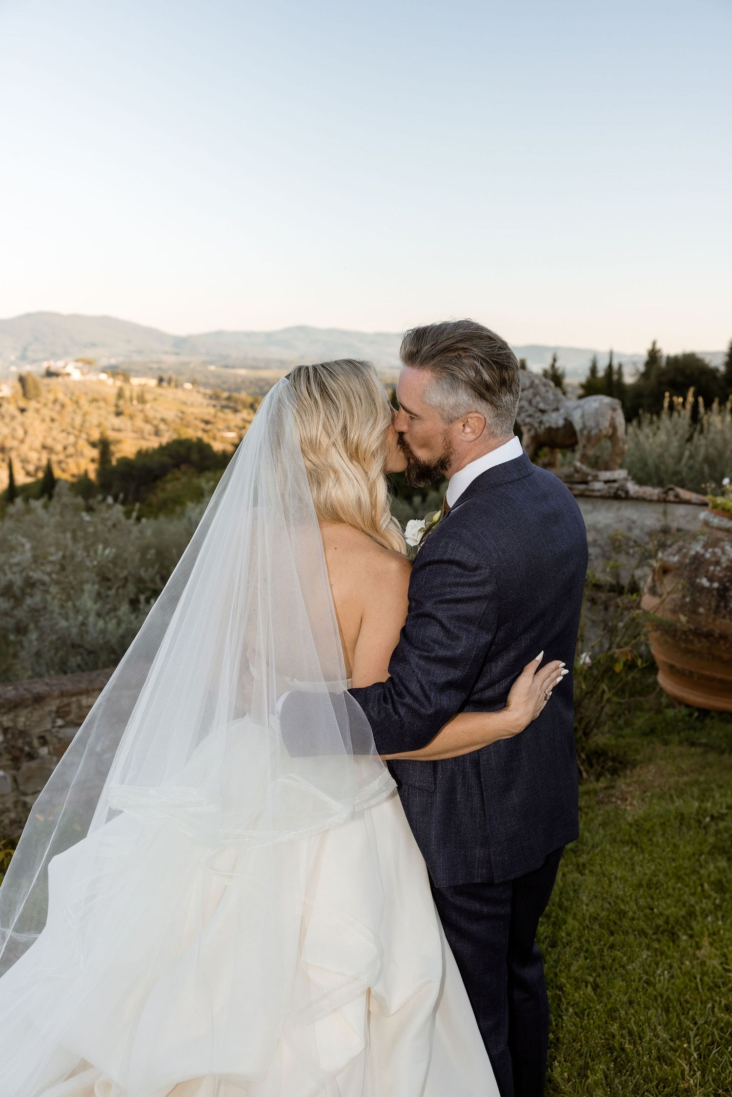 Newlywed couple sharing romantic kisses against scenic Tuscan countryside backdrop at sunset.