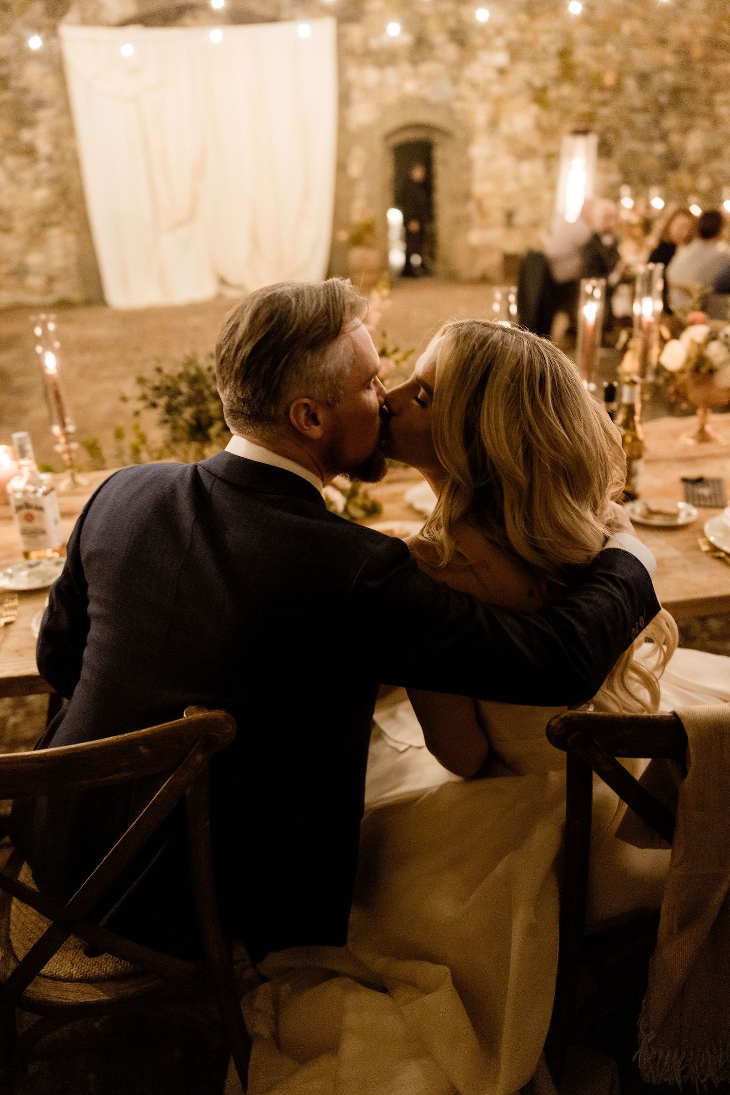 A couple sits intimately at an elegant candlelit dinner table in a rustic stone-walled venue during a romantic evening.
