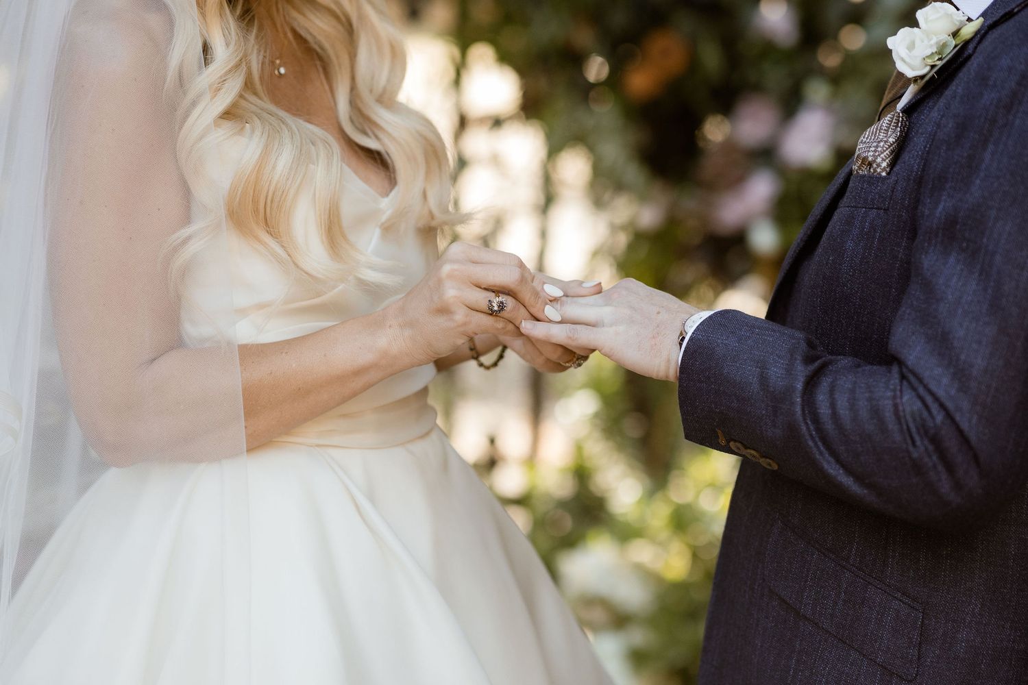 A couple holds hands during an outdoor wedding ceremony with sunlight filtering through trees in the background.