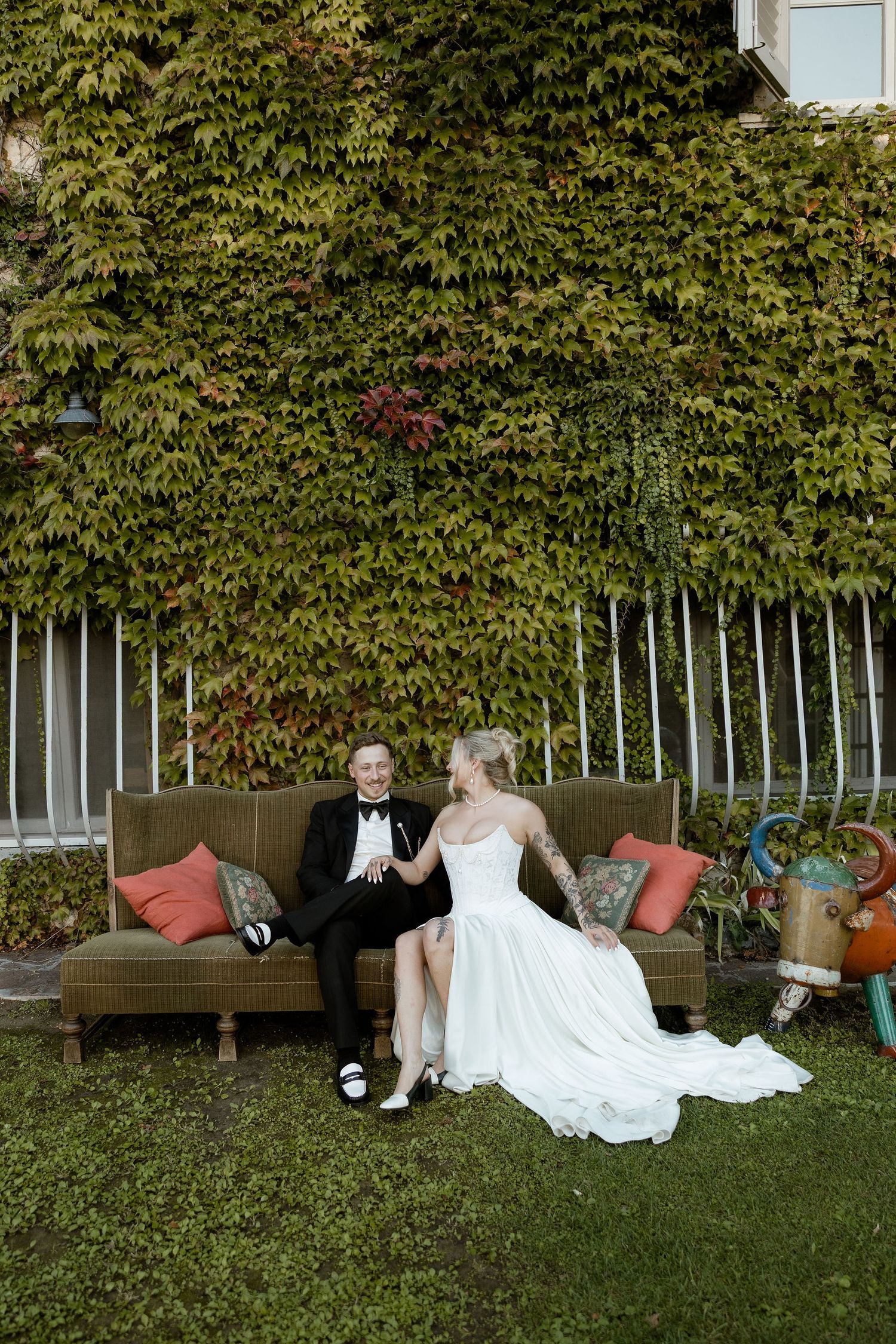 A couple in wedding attire sits on a bench against an ivy-covered wall in a garden setting.