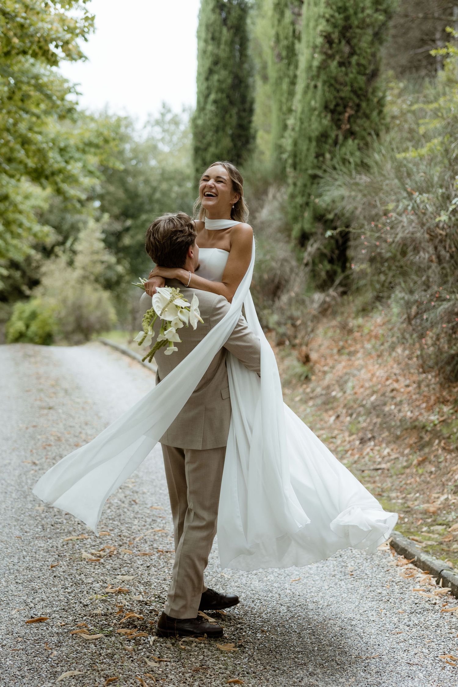 A joyful wedding couple shares a playful moment on a tree-lined gravel path in an outdoor setting.