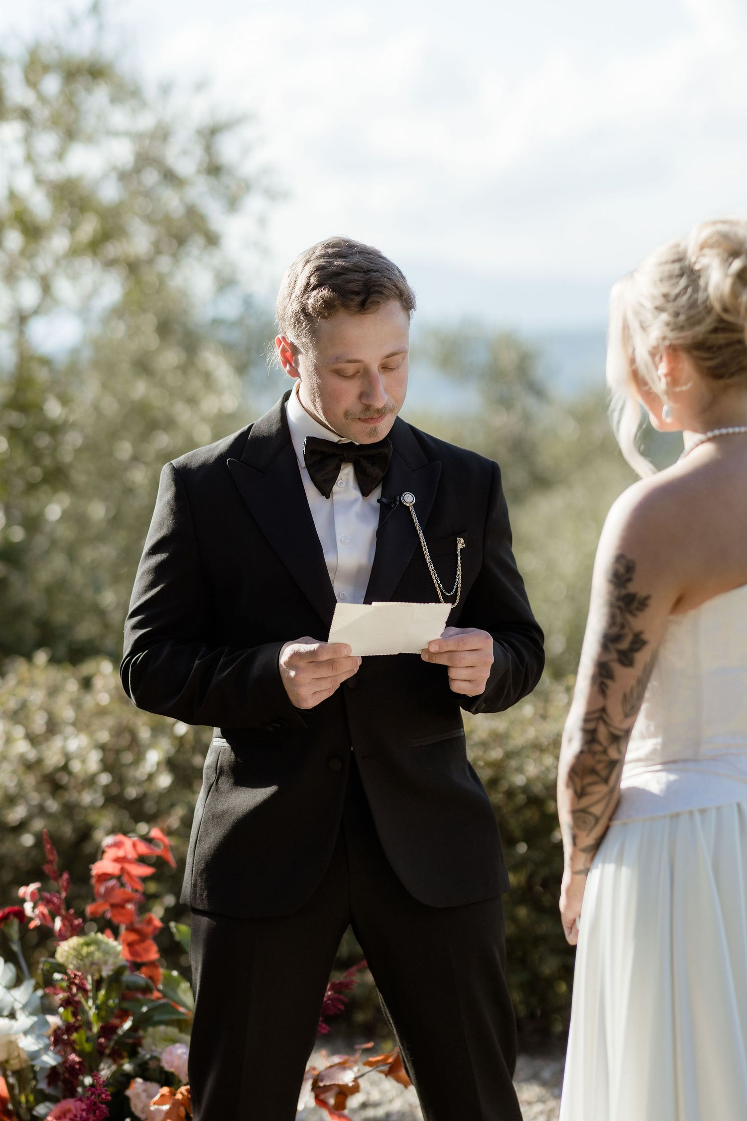A groom reads wedding vows from a paper during an outdoor mountain ceremony with red flowers in the background.