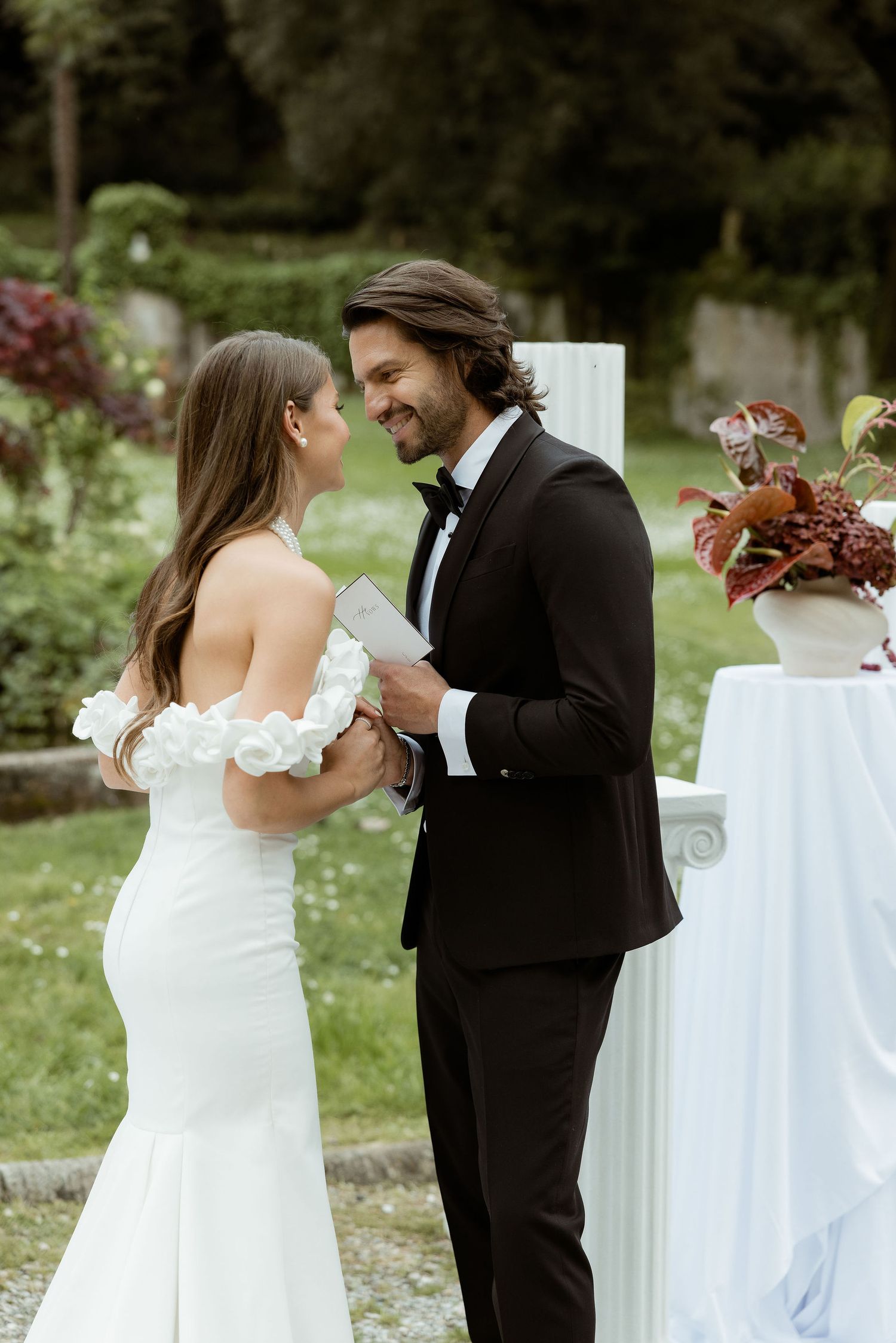 A romantic outdoor wedding scene with a couple reading vows in a garden setting with floral decor and white table accents.