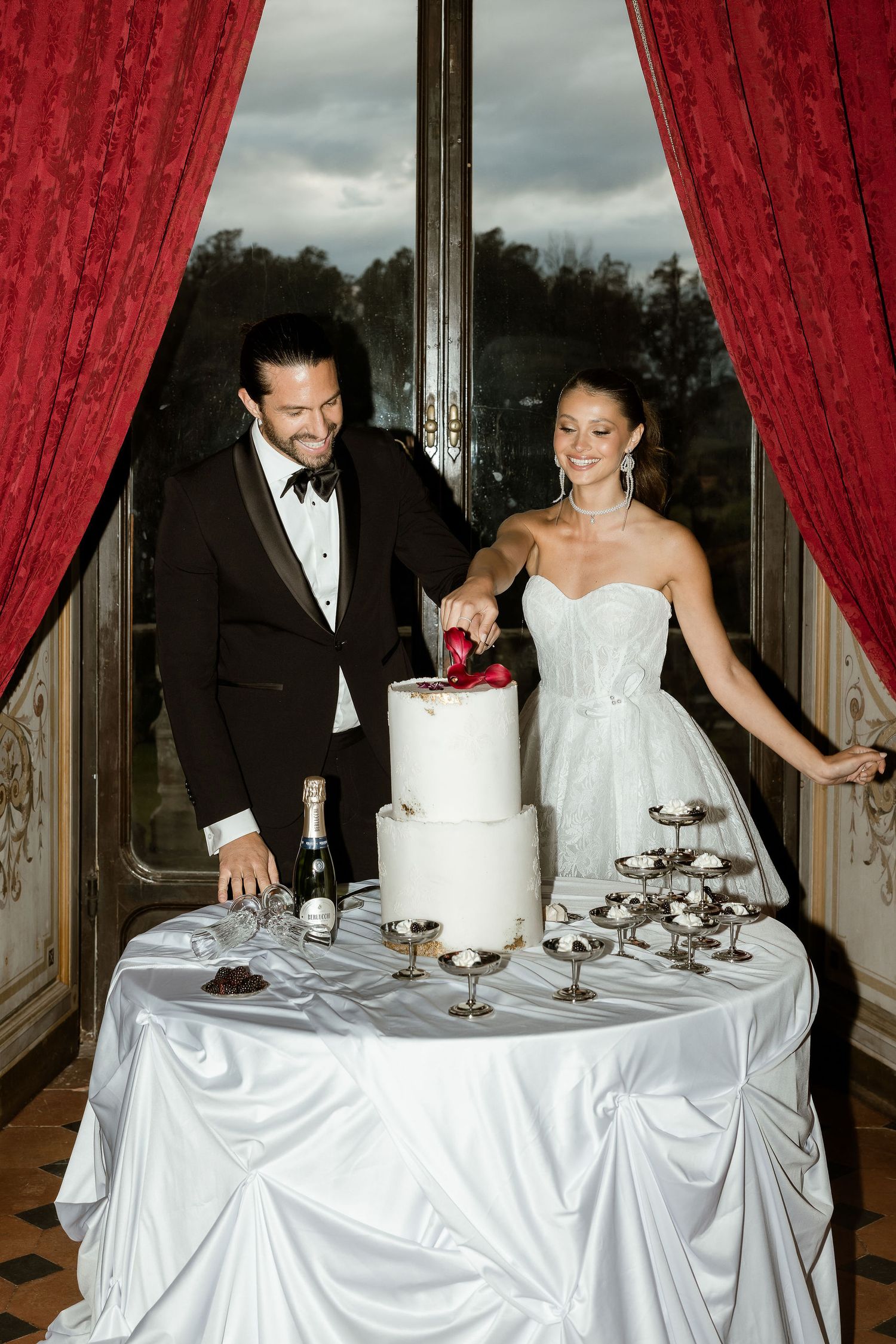 A couple in formal attire cut their elegant white wedding cake beside crimson curtains at an indoor reception.