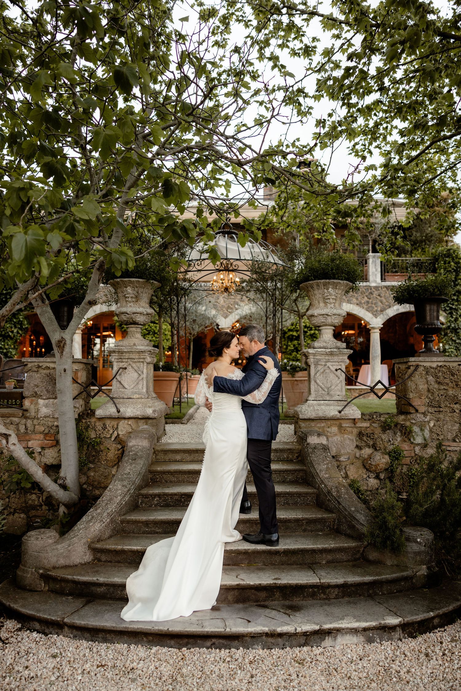 A couple embraces on an elegant stone staircase surrounded by lush greenery and archways at an outdoor garden venue.