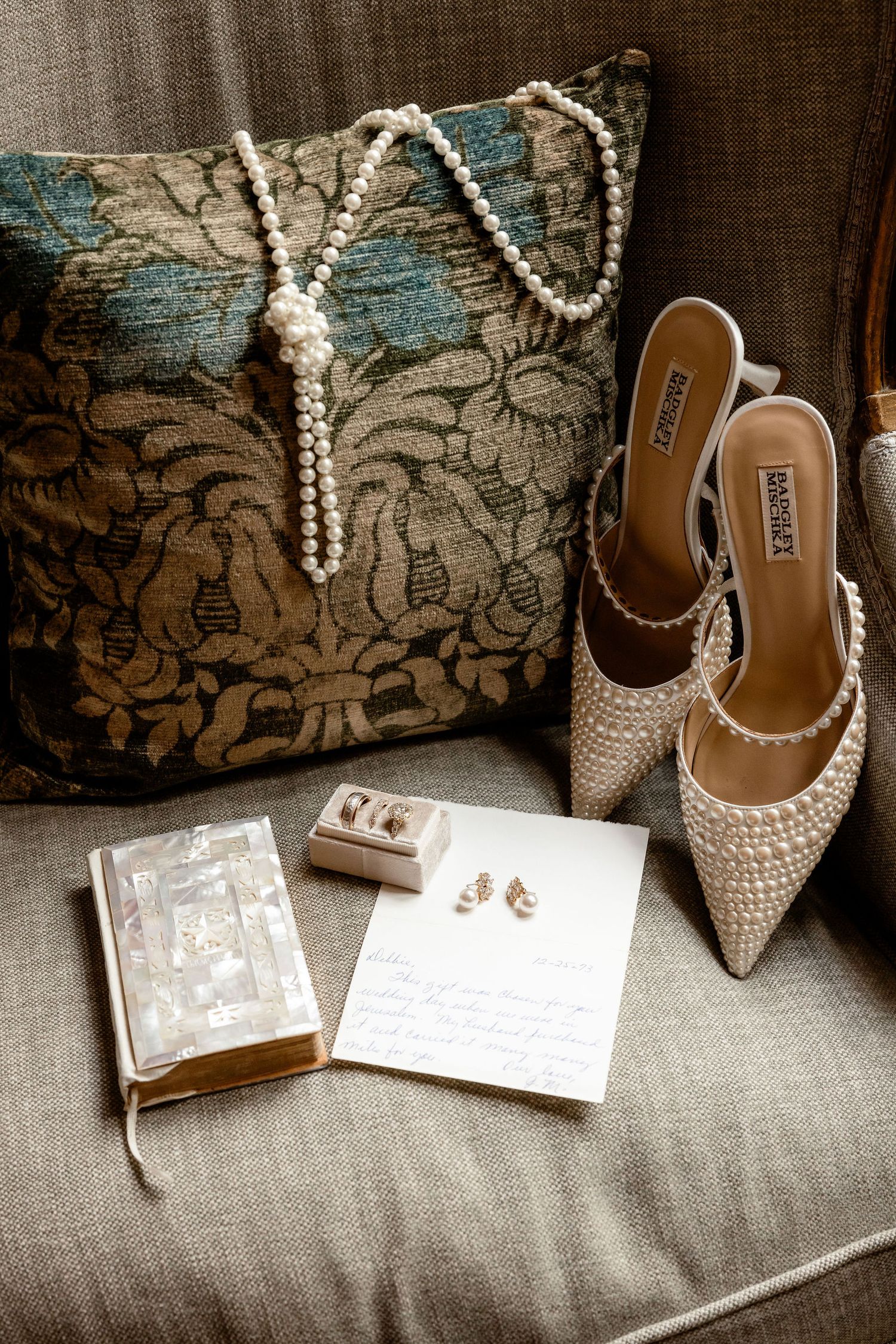 Wedding accessories including pearl necklace, beaded shoes, and decorative pillow arranged with invitation on gray couch.