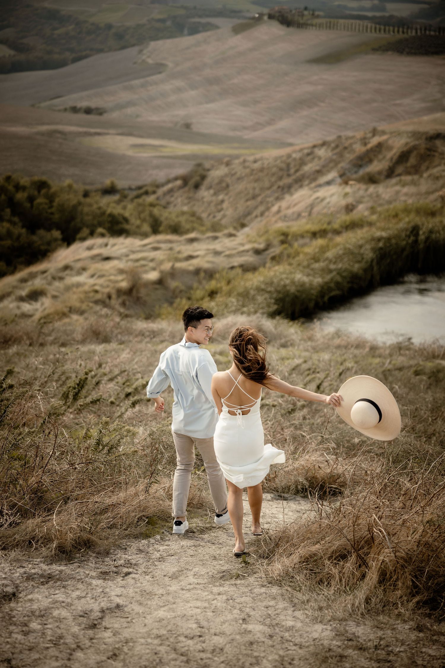 A couple walking hand in hand on a hillside path with a straw hat caught in the wind.