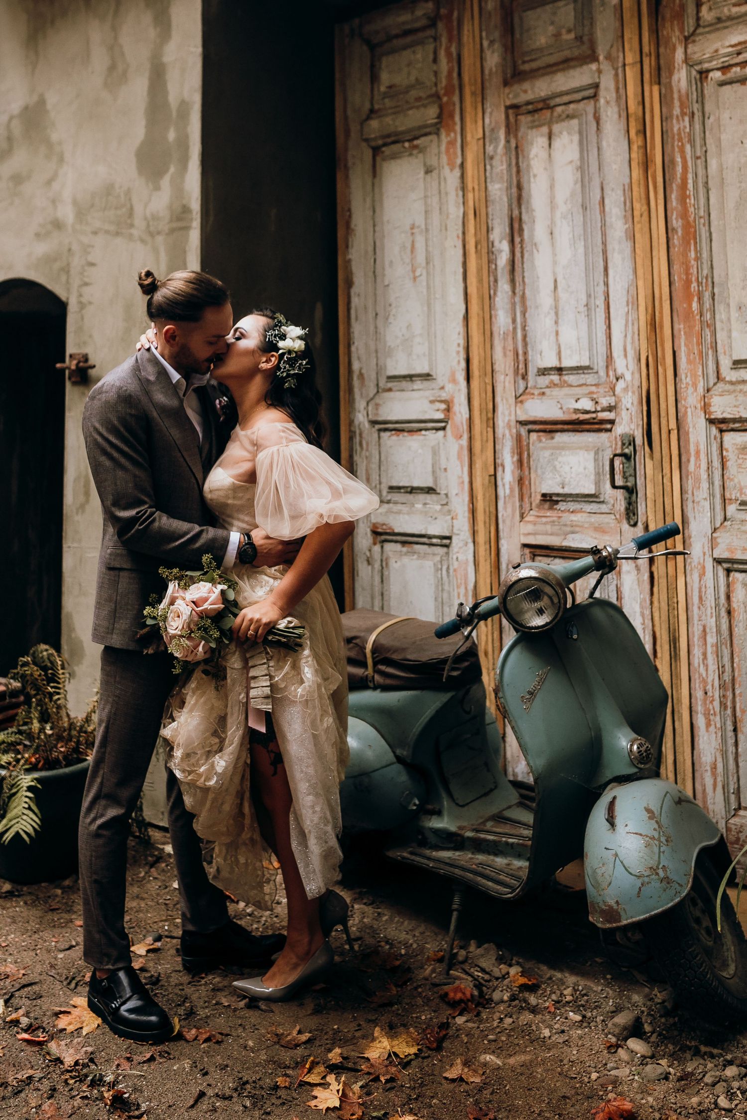 A romantic vintage-styled wedding couple poses beside a classic green Vespa scooter against rustic wooden doors.