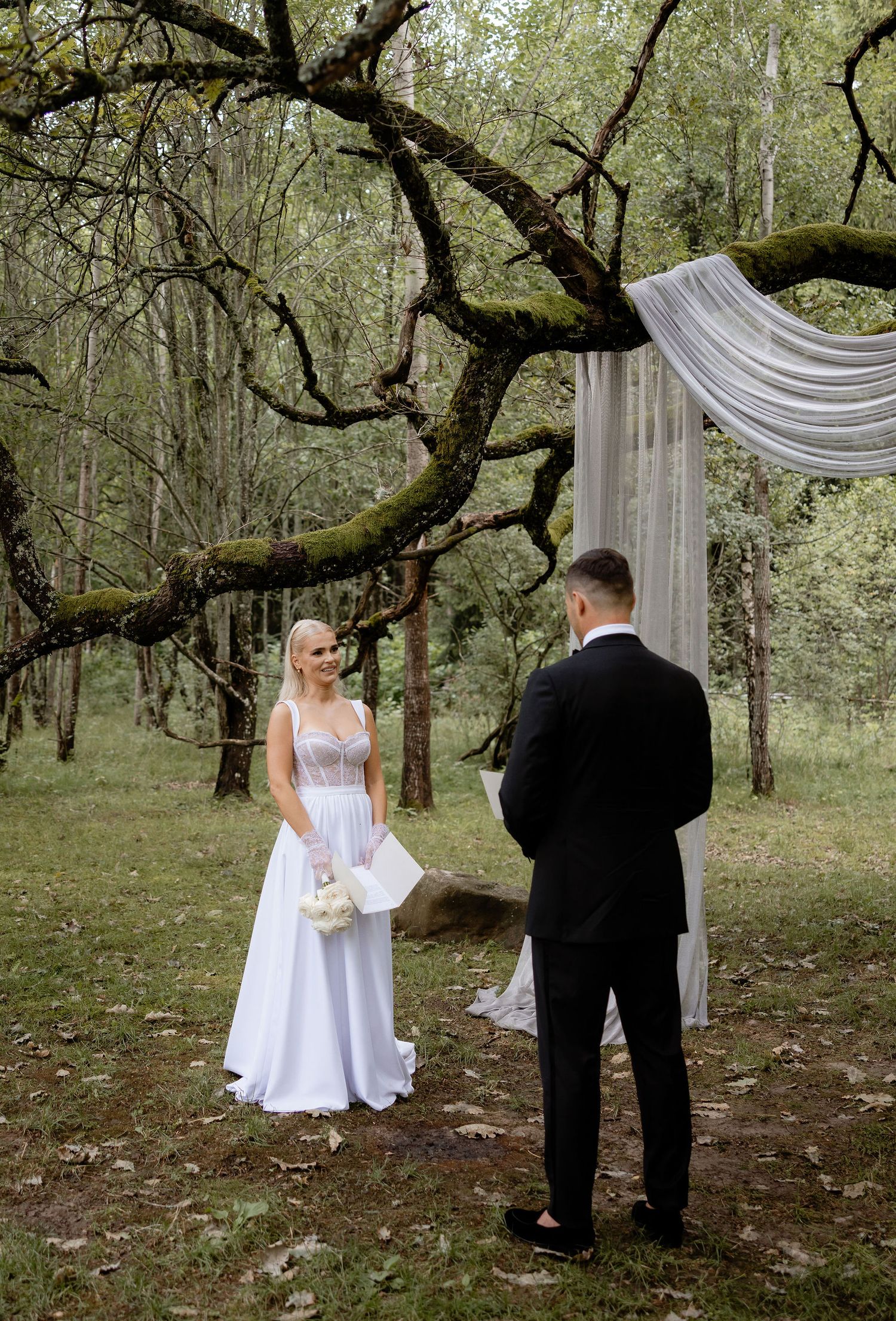 An intimate forest wedding ceremony with white draped fabric arch and moss-covered trees.