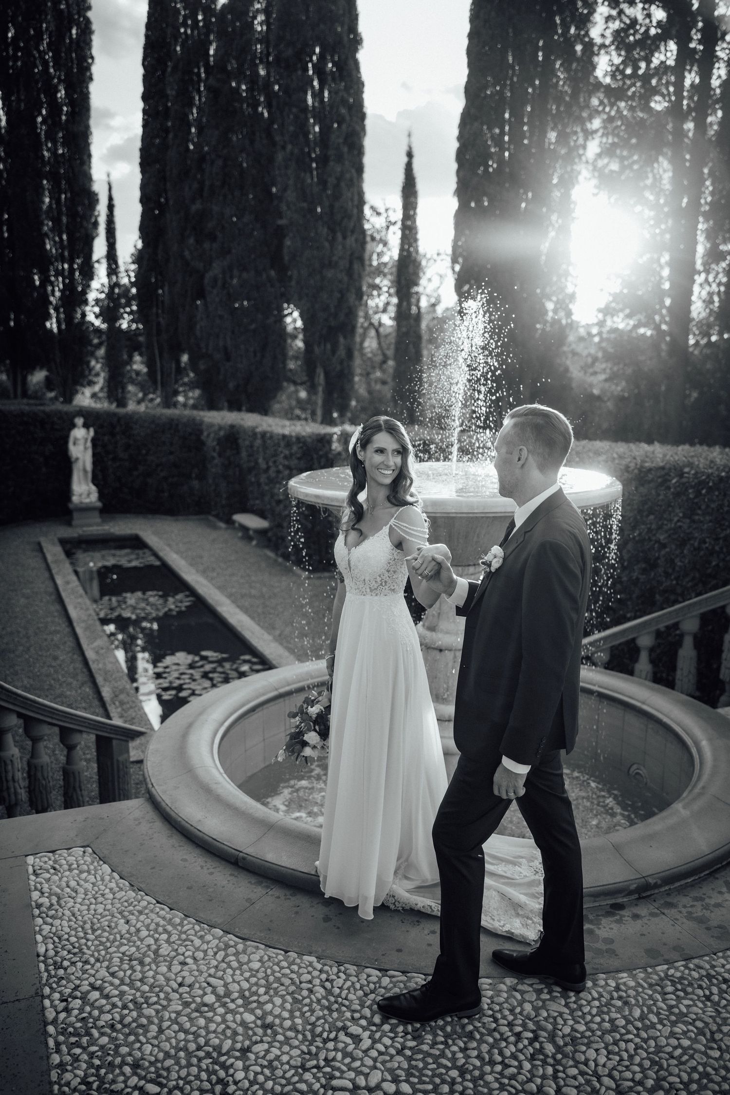 A newlywed couple poses by an ornate fountain in a garden with sunlight streaming through cypress trees.