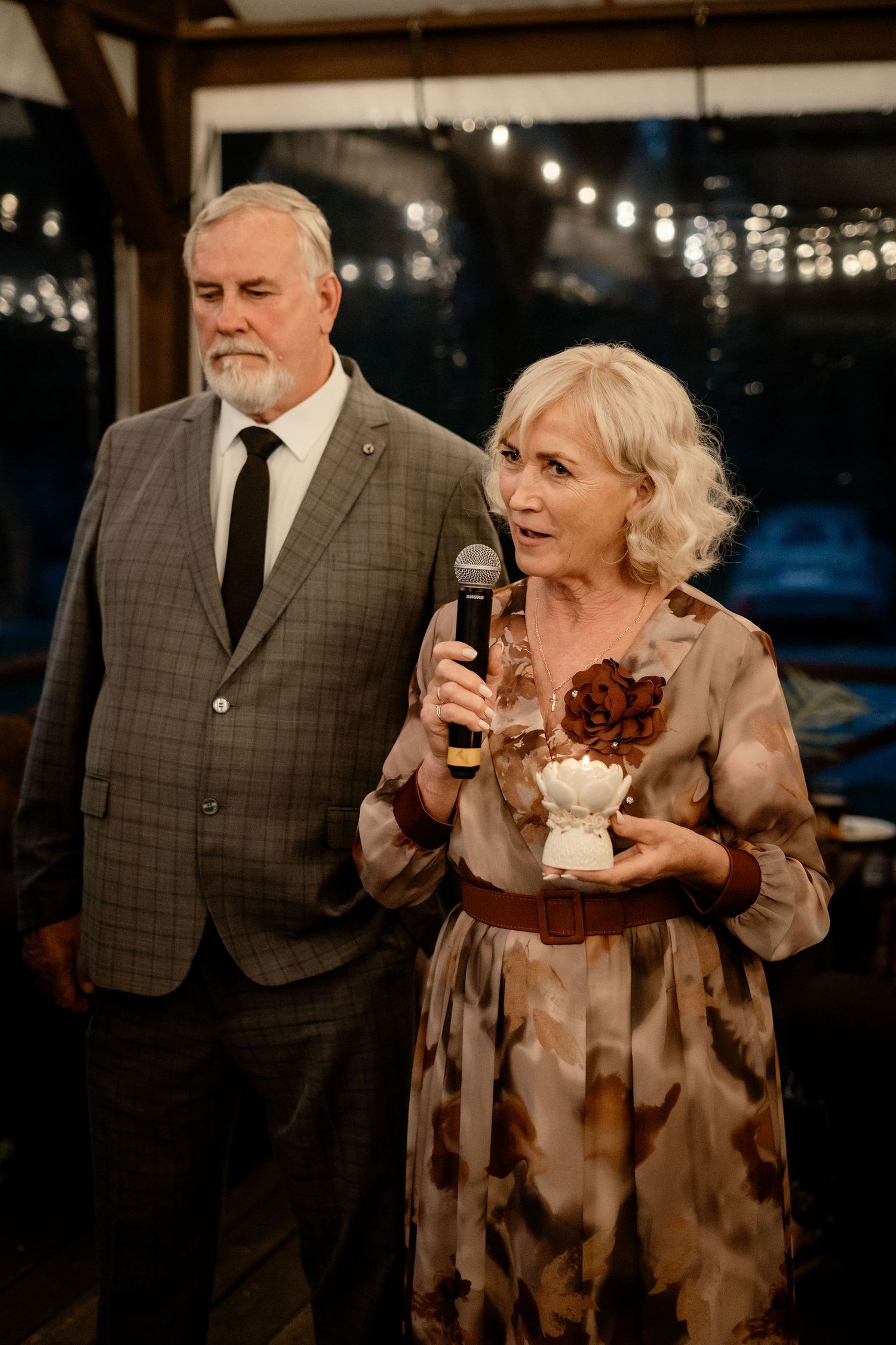 A couple speaks at an evening event, with the woman holding a microphone and a small item while wearing a brown patterned dress.