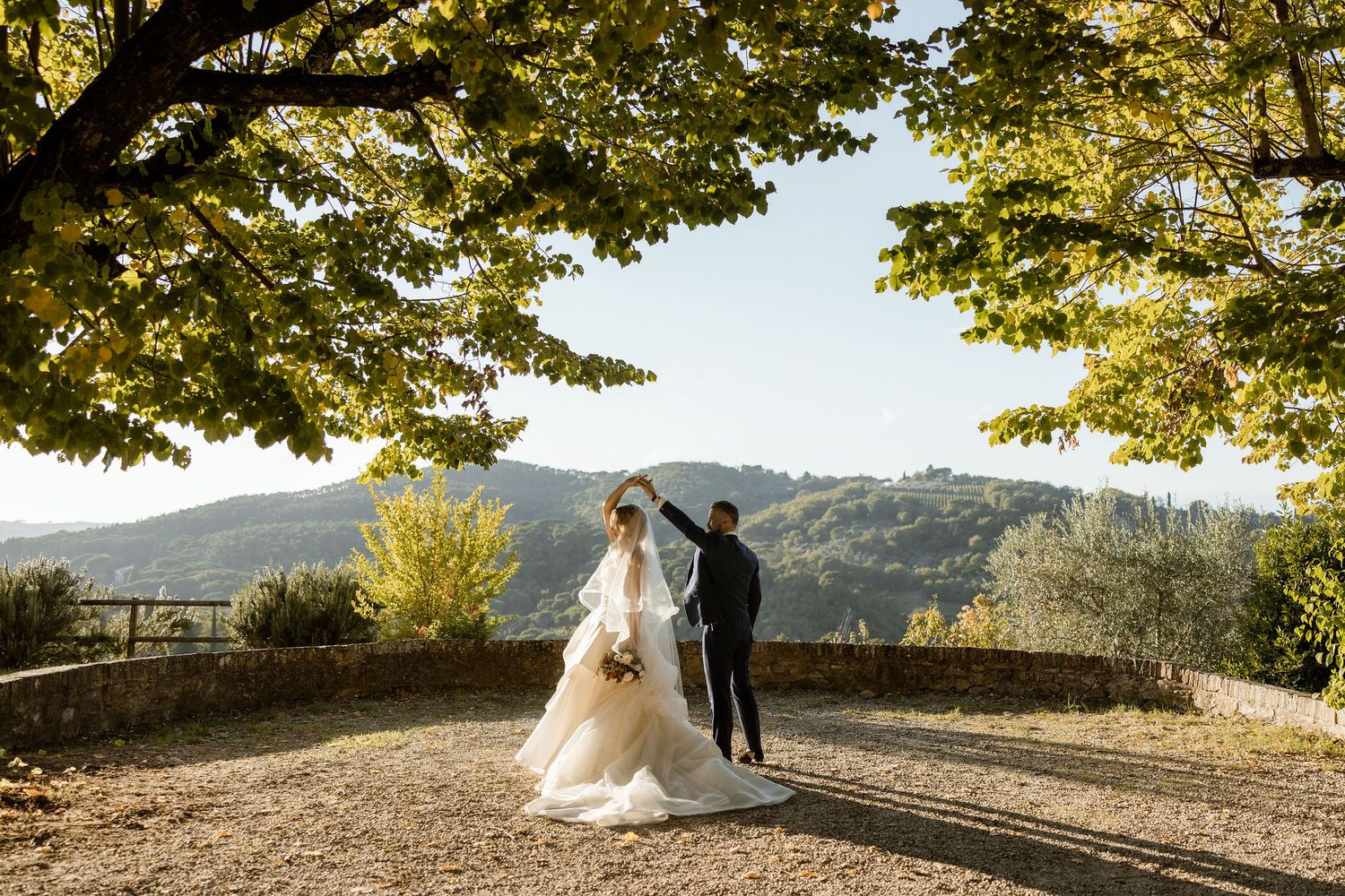 A scenic outdoor wedding photoshoot with a couple on a stone wall overlooking a mountainous landscape in autumn.